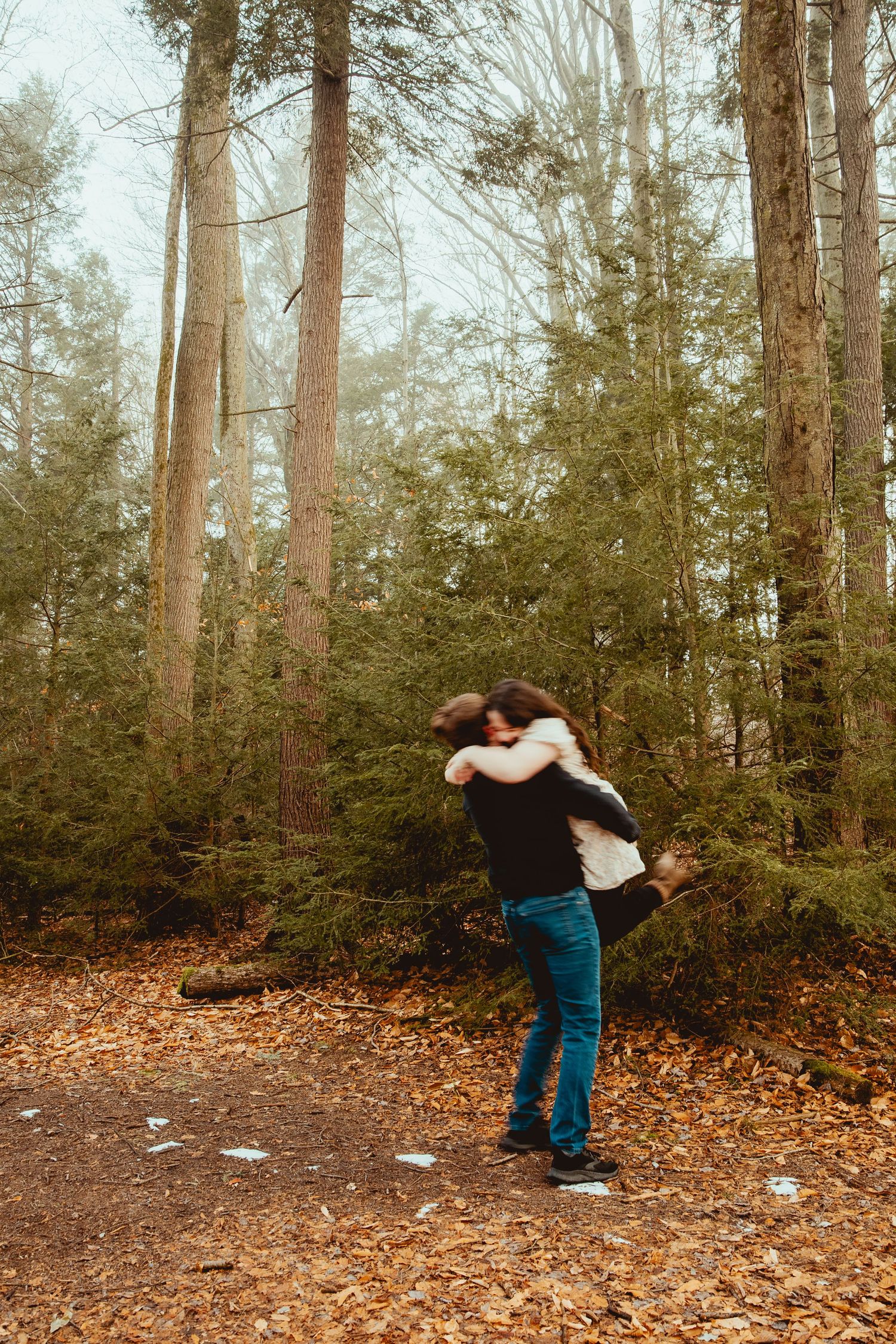 Two people share an intimate embrace surrounded by tall evergreen trees and autumn foliage.