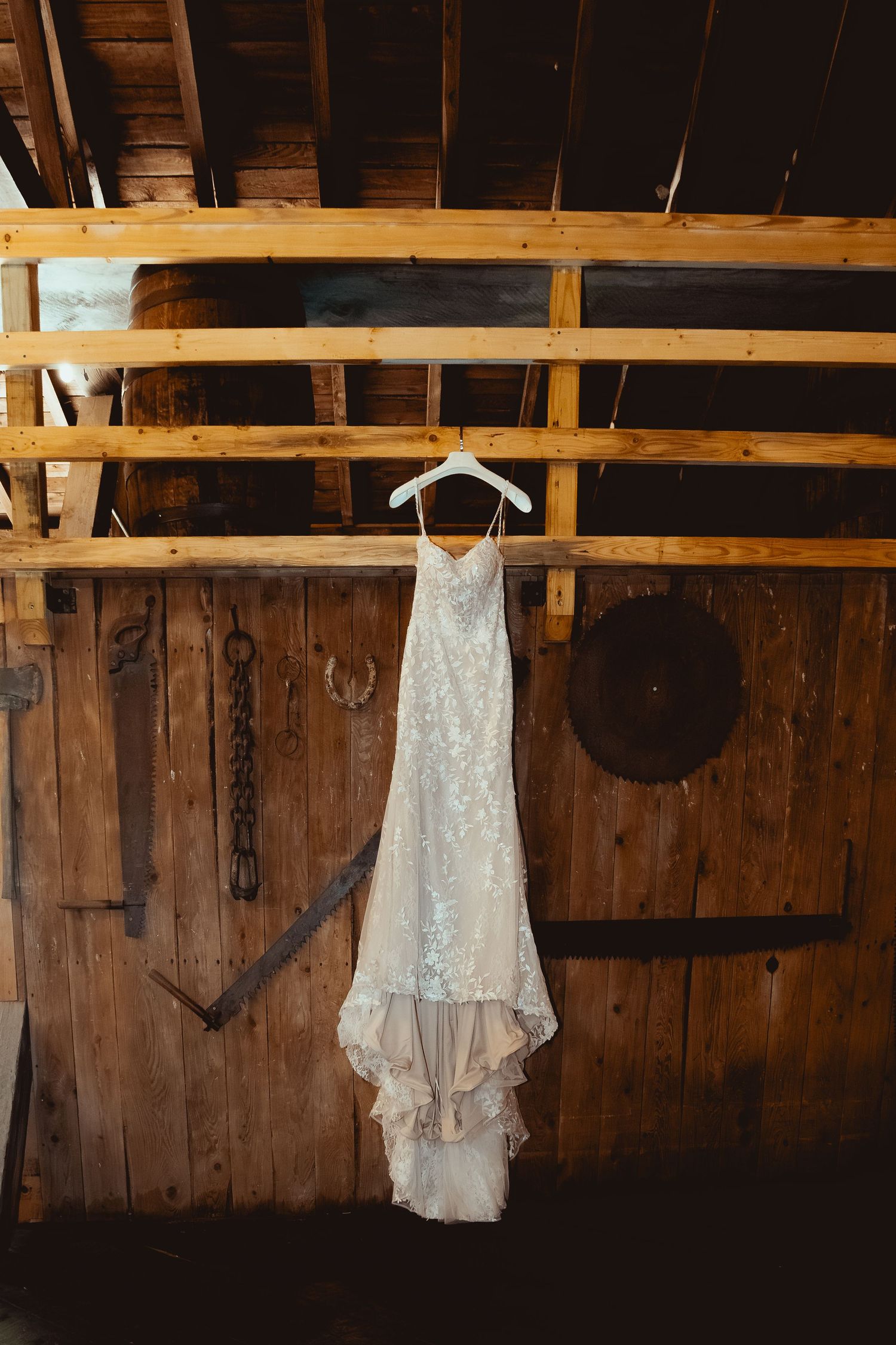 White lace wedding dress hanging on wooden beams in rustic barn setting with dramatic lighting.
