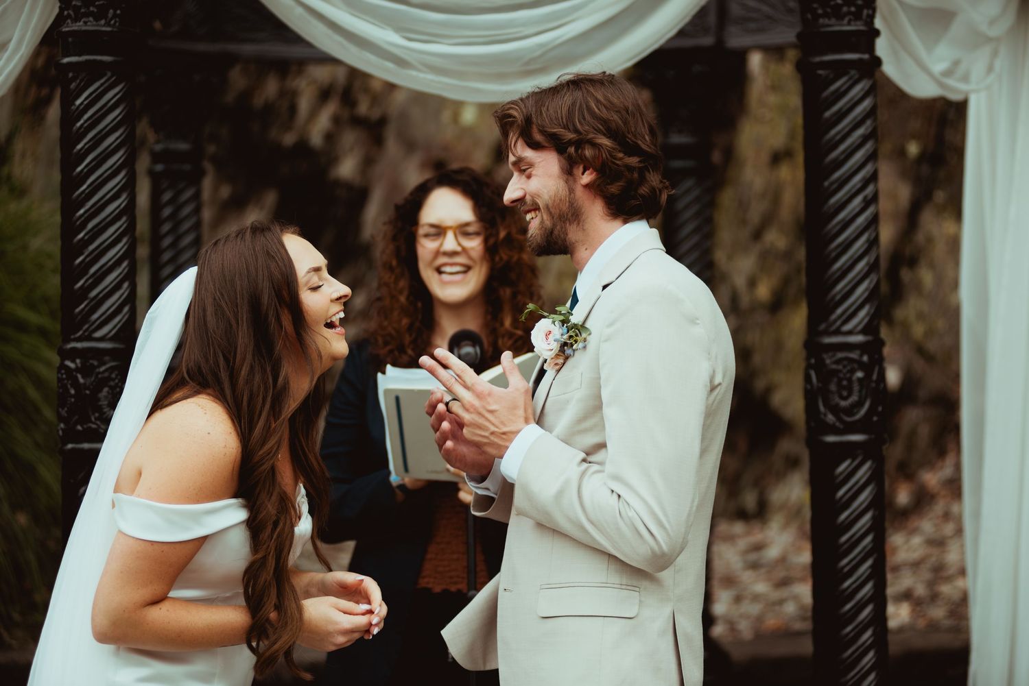 Bride and groom laughing with officiant during outdoor wedding ceremony under decorative arch.