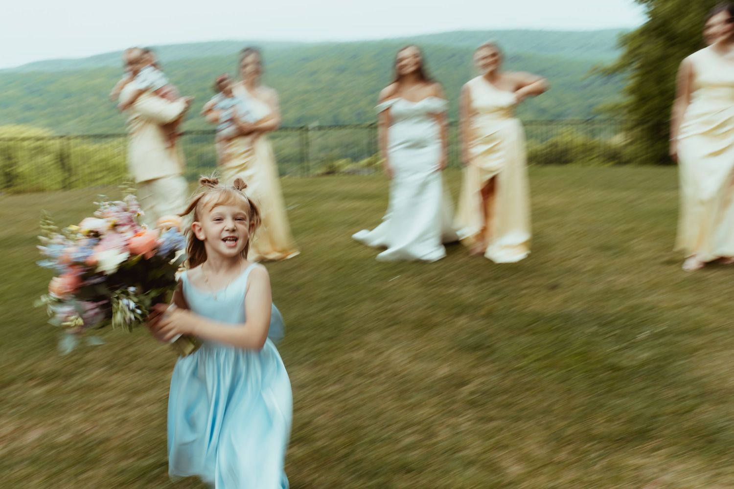 Joyful flower girl in blue dress running with bouquet, bridesmaids in yellow gowns on green hillside.