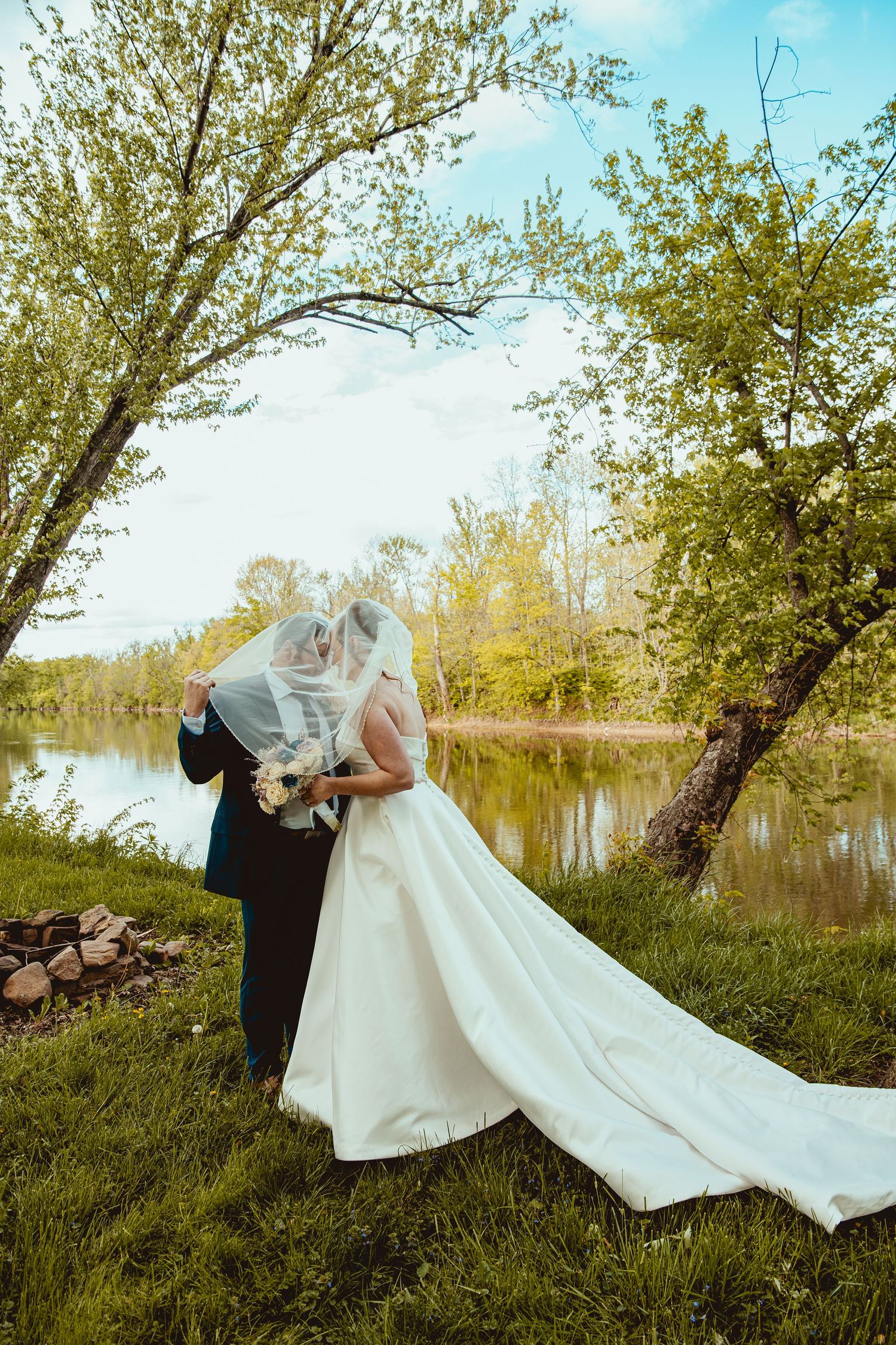 Bride and groom share a kiss under veil beside a serene lake in lush greenery.