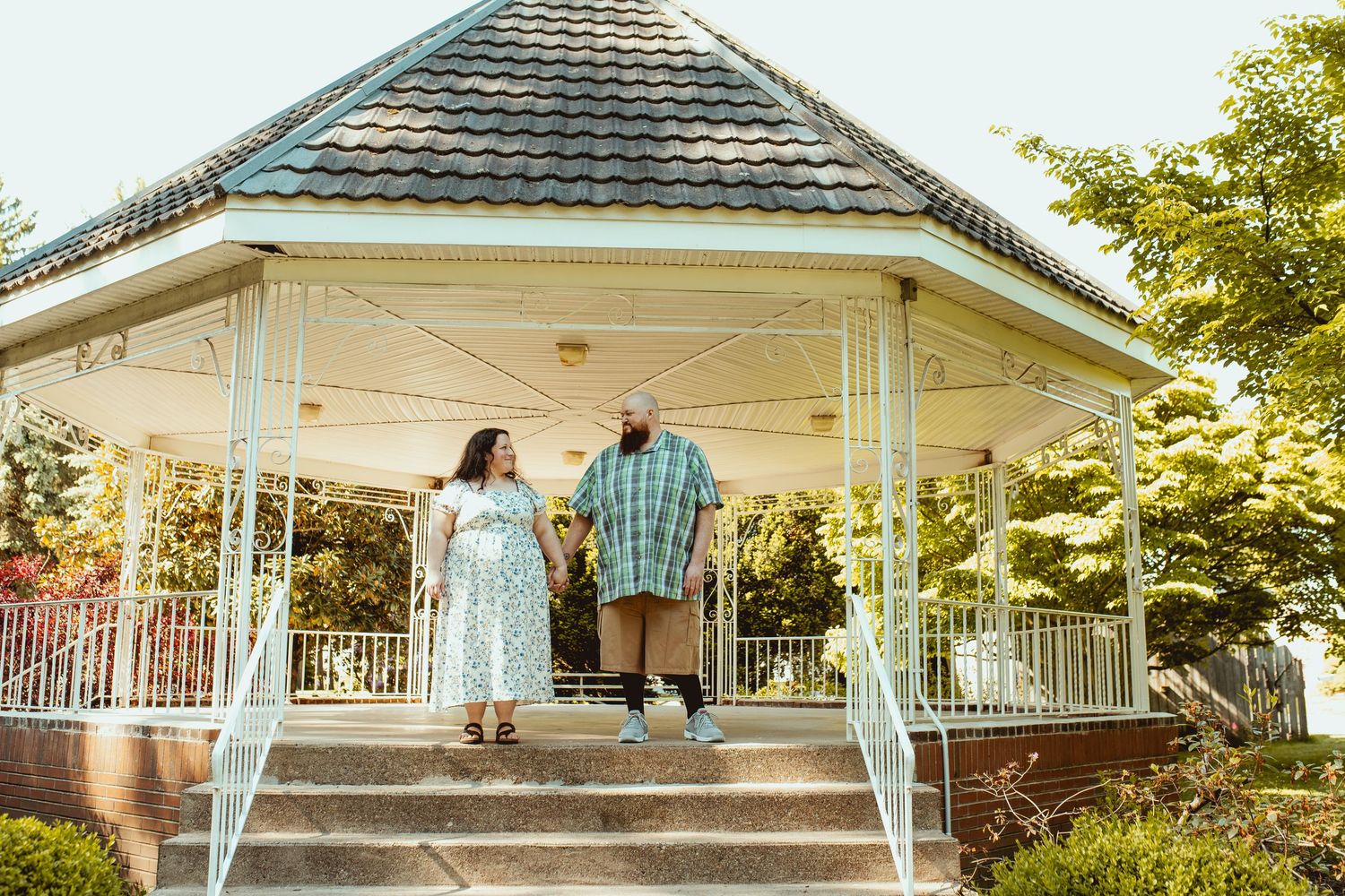 Couple holding hands on steps of a white gazebo with green roof surrounded by trees and gardens.