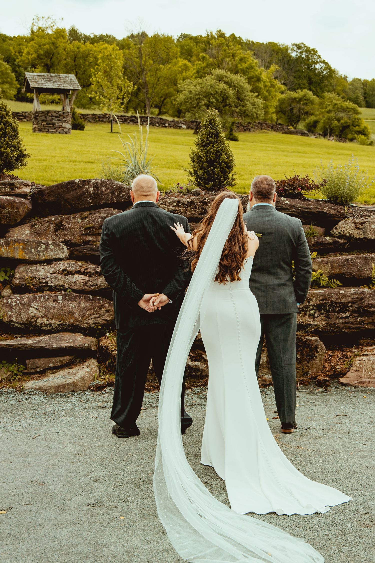 Back view of a bride in white gown with two men in formal suits standing outdoors near stone wall.