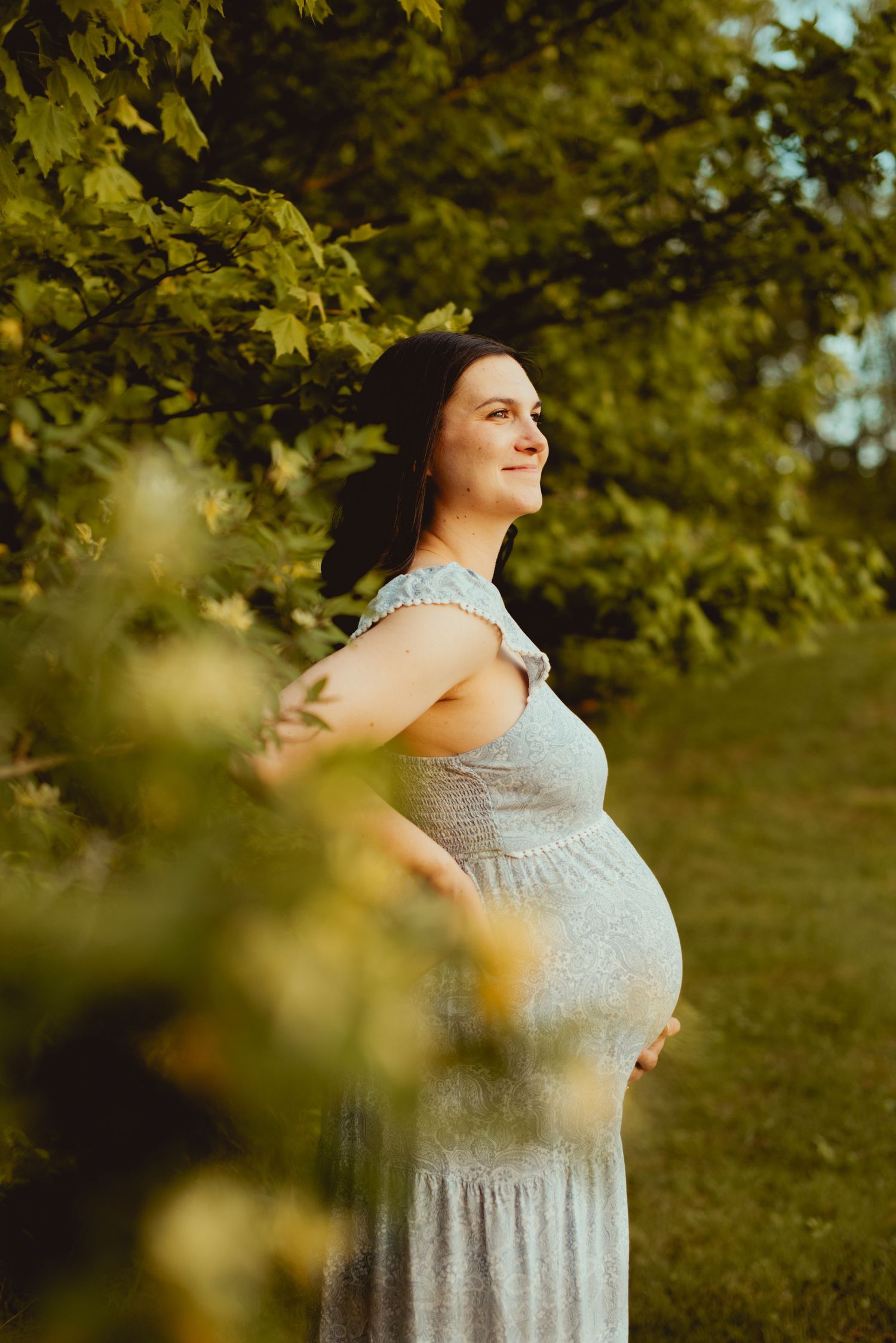 A pregnant woman in a white floral dress stands among lush green trees during a summer photoshoot.