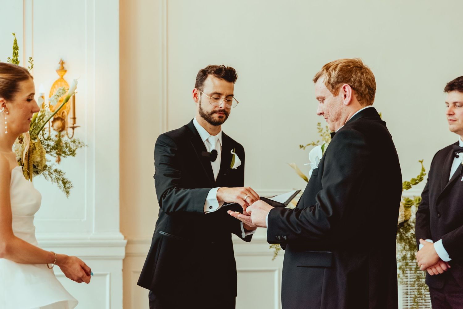 Wedding party members in formal attire interact during ceremony in elegant indoor venue.