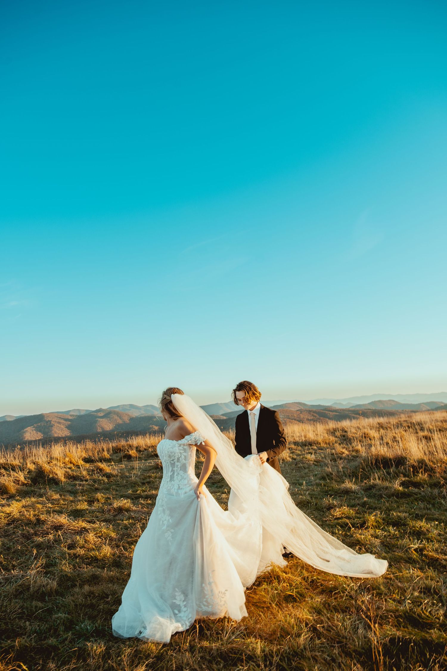 Bride's white dress and veil flowing in the wind against mountain landscape at sunset.
