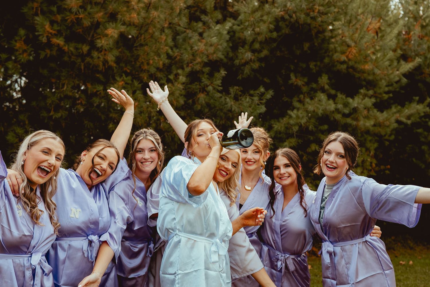 A group in matching purple and blue robes laugh and celebrate together outdoors during a wedding celebration.