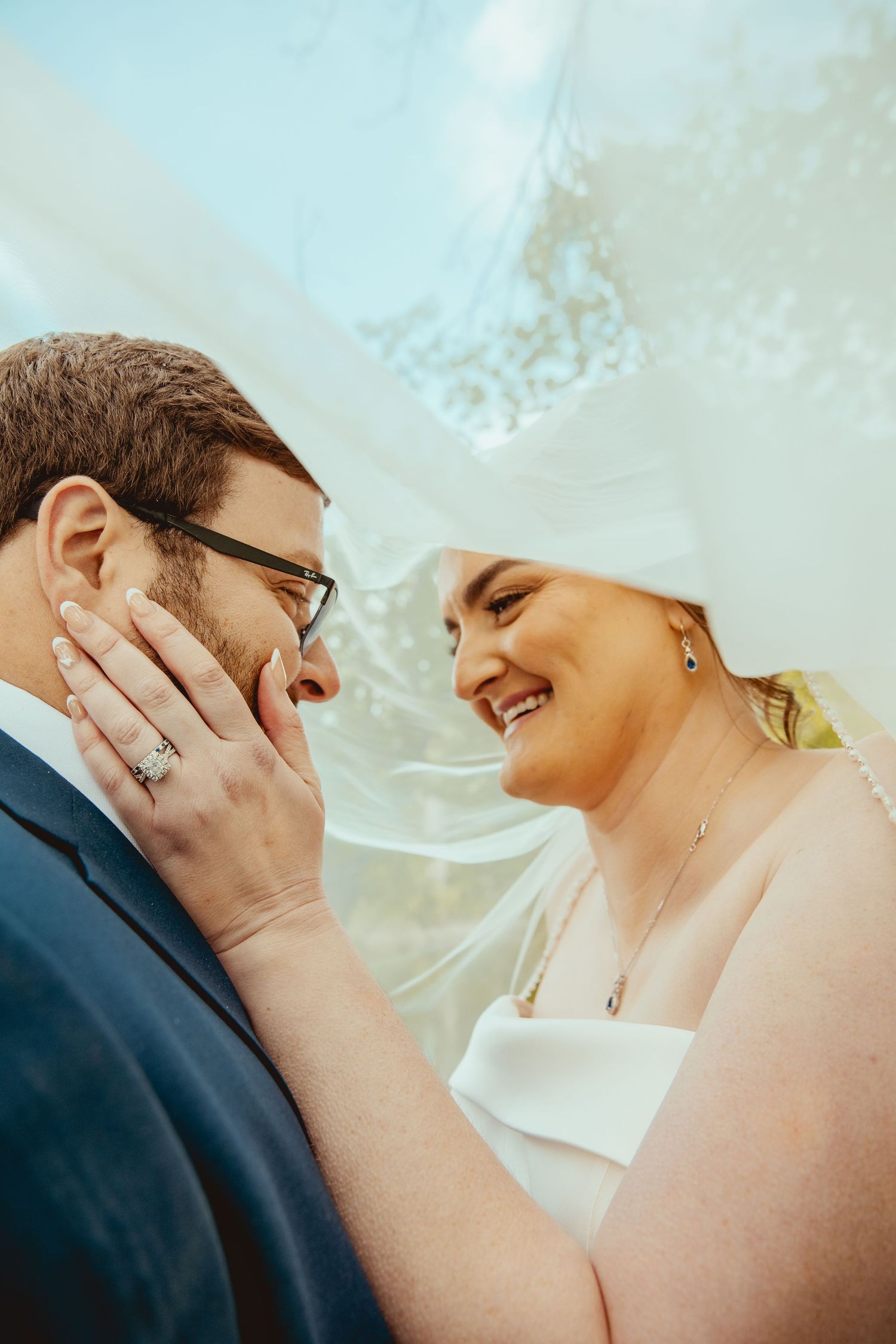 Joyful bride and groom sharing intimate moment under wedding veil, bride touching grooms face with diamond ring.