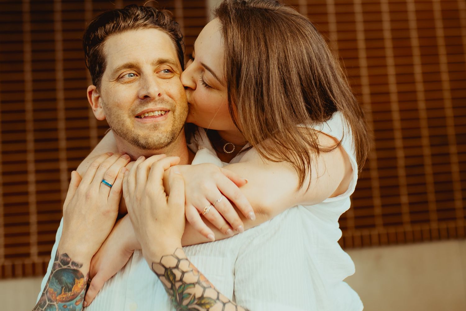 A tattooed man smiles as a woman kisses his cheek, embracing him warmly in a cozy outdoor setting.