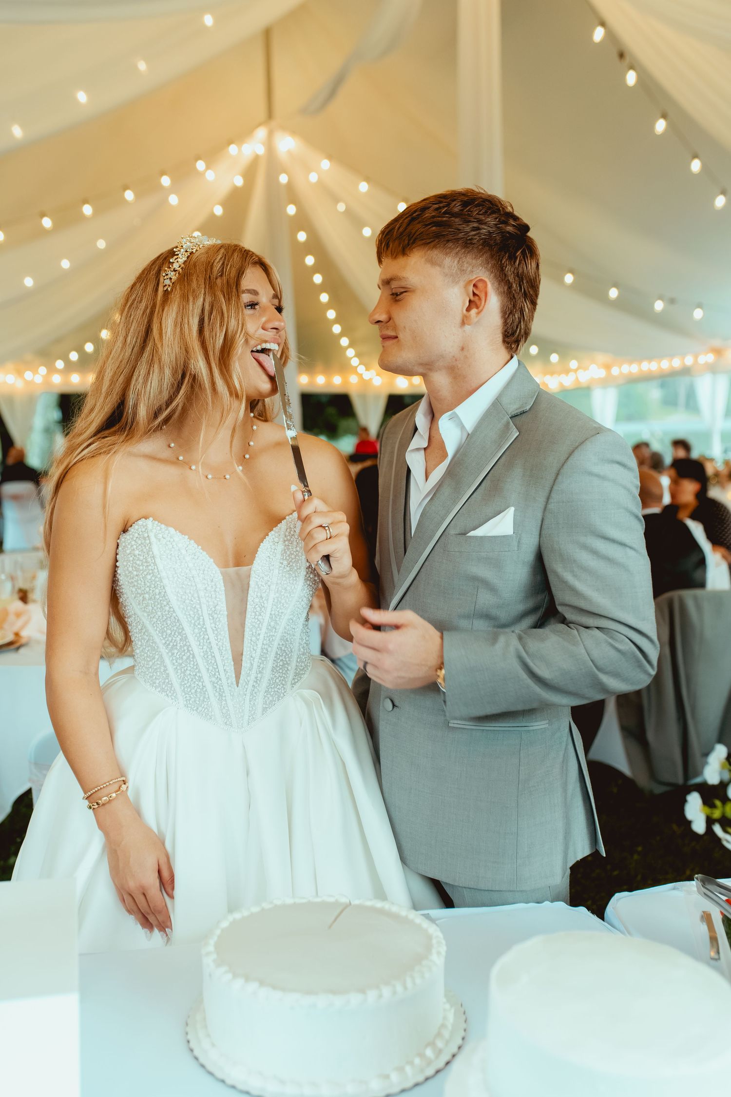Bride and groom cutting white wedding cake together under string lights at reception.