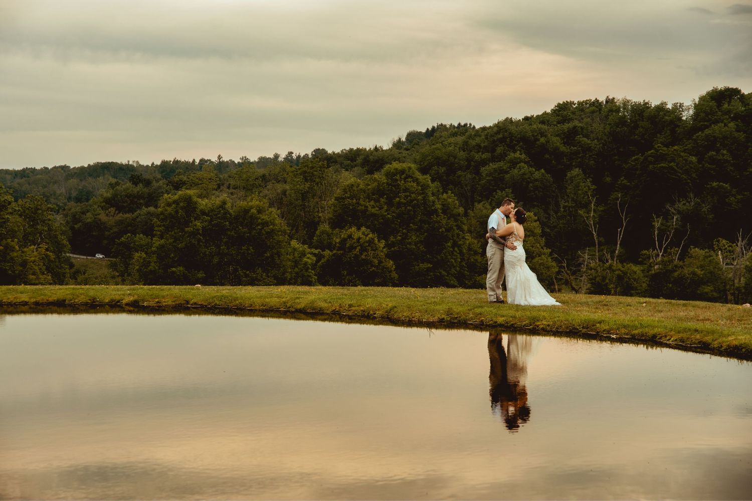 Bride and groom share romantic kiss reflected in still pond at golden hour sunset.
