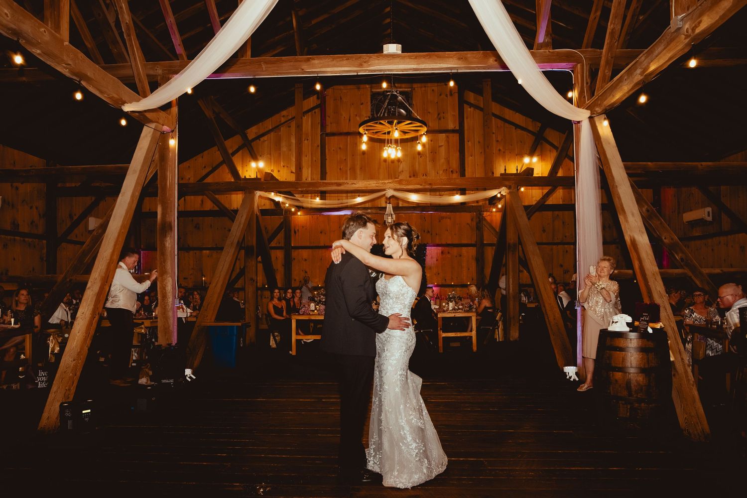 First dance in rustic barn reception with romantic lighting and wooden beams overhead.