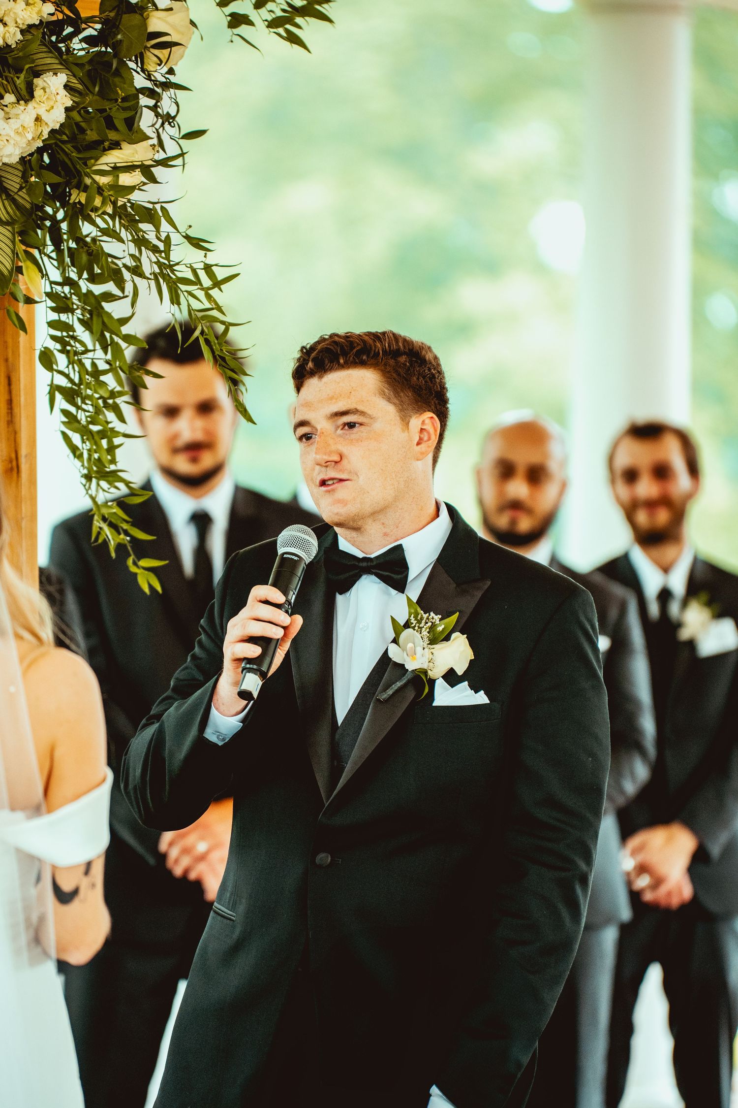 Groomsmen in black tuxedos gather during wedding speeches at an elegant indoor venue.