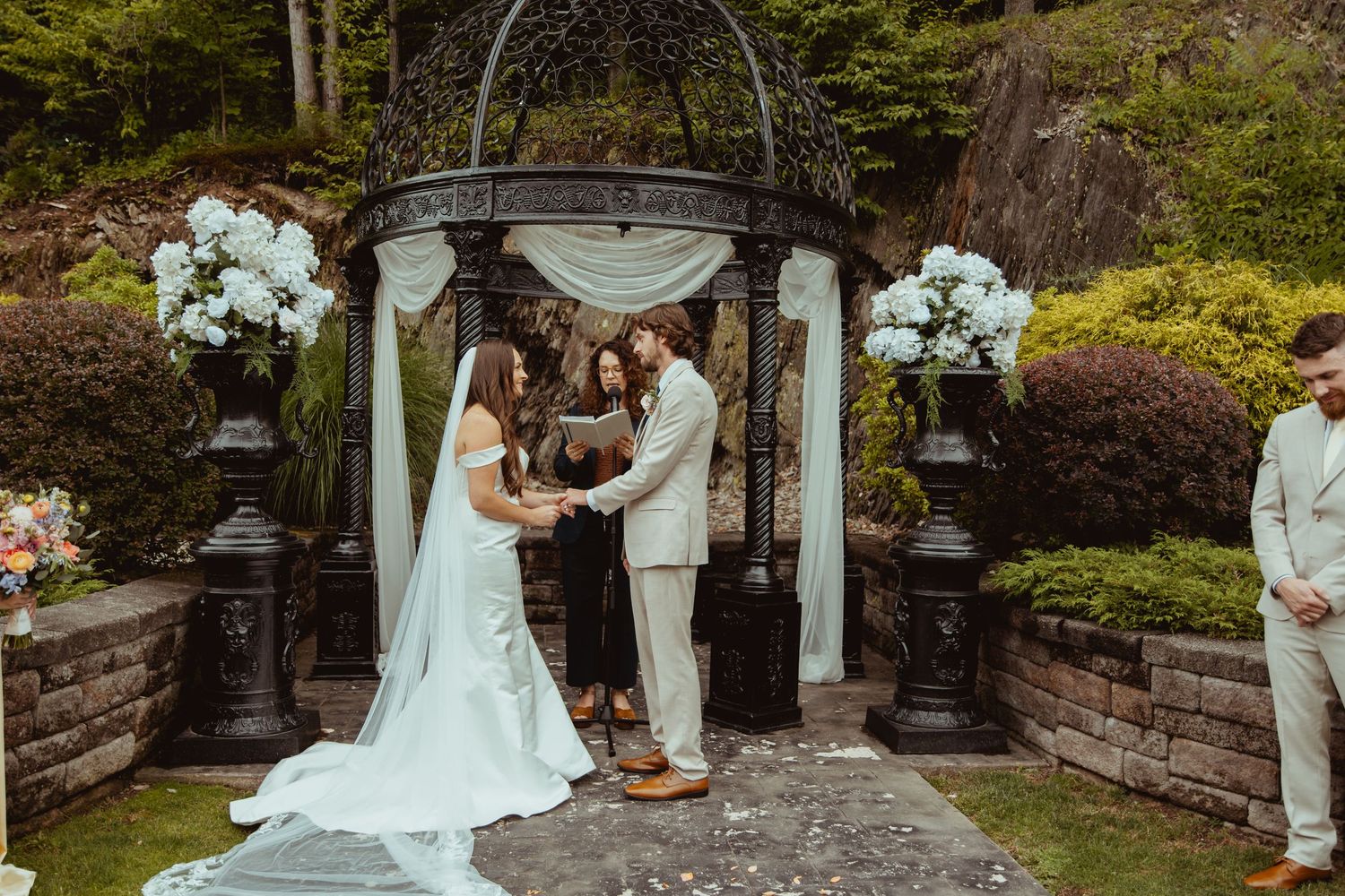 Bride and groom exchange vows under an elegant outdoor gazebo adorned with white hydrangeas during a garden wedding ceremony.