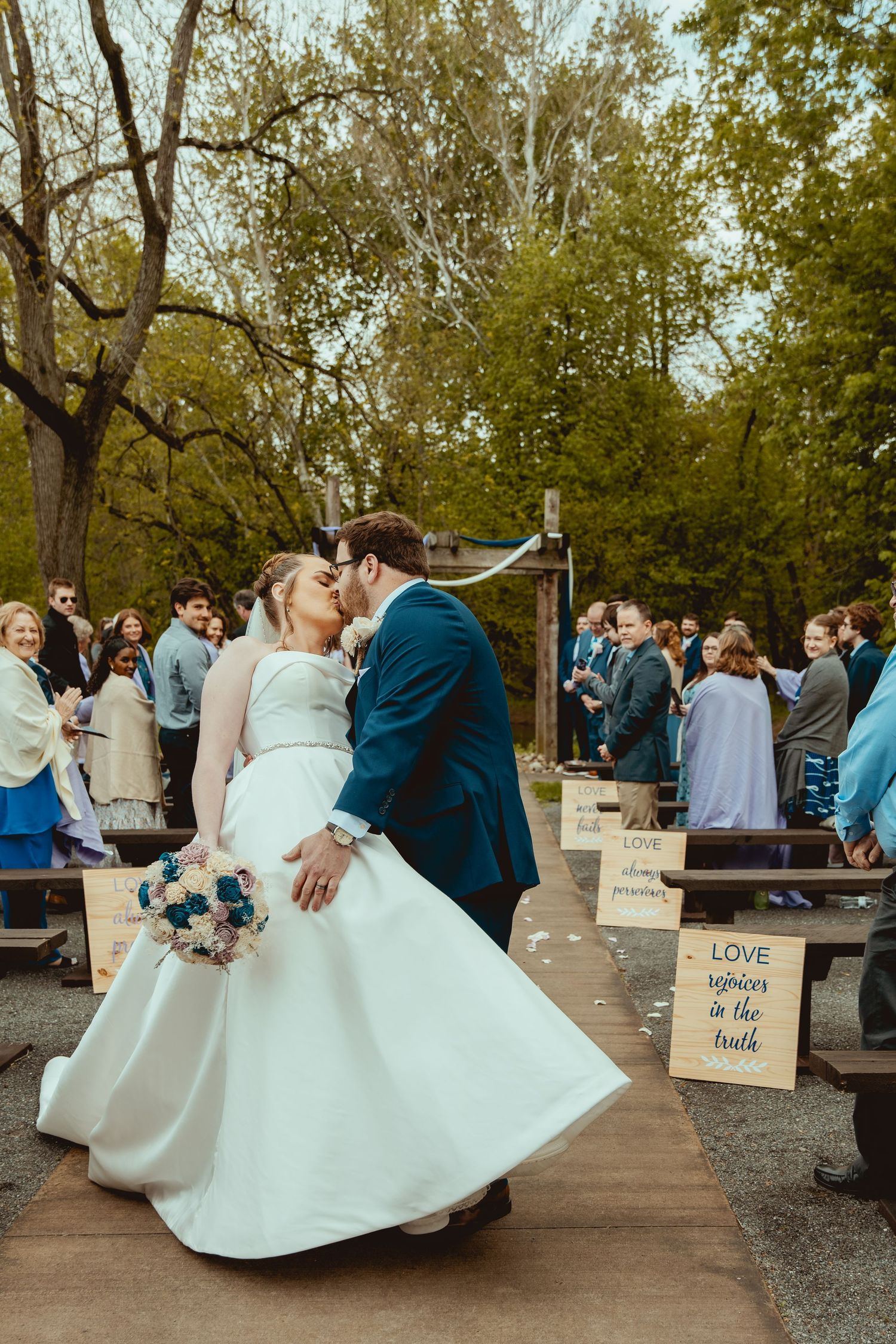 Newlyweds share a romantic first kiss as husband and wife along an aisle lined with wooden signs at outdoor wedding.