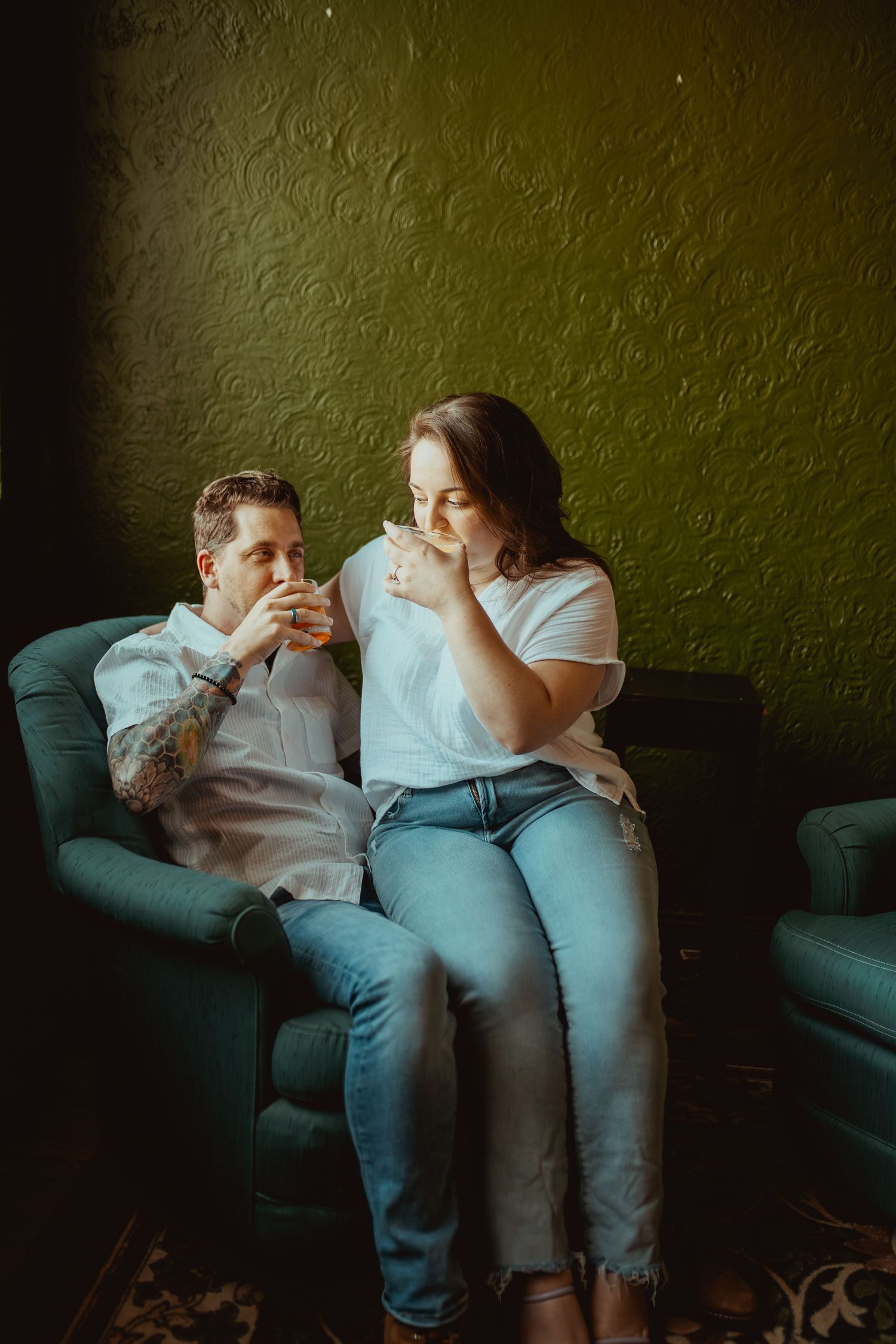 A couple sitting together on a teal armchair, sharing drinks against a green textured wall.