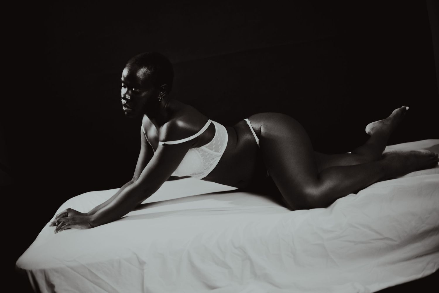 Black and white photo of a woman in white lingerie posing gracefully on a white bed against a dark background.