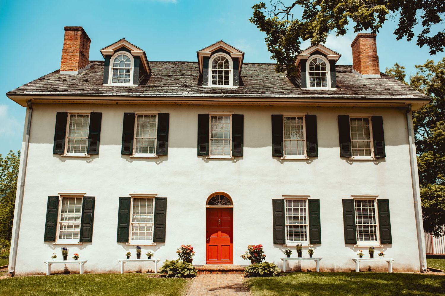 Historic white colonial house with black shutters, red door, and three dormer windows stands against a blue sky.
