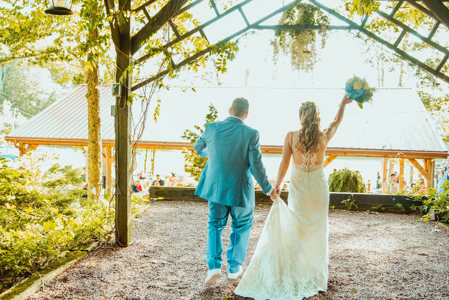 Newlywed couple walking along a scenic outdoor path surrounded by trees and sunlight.