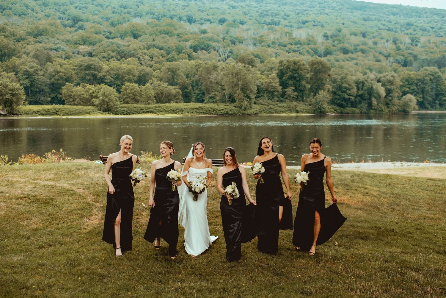 Wedding party poses together on grassy hillside overlooking scenic lake and mountain landscape.