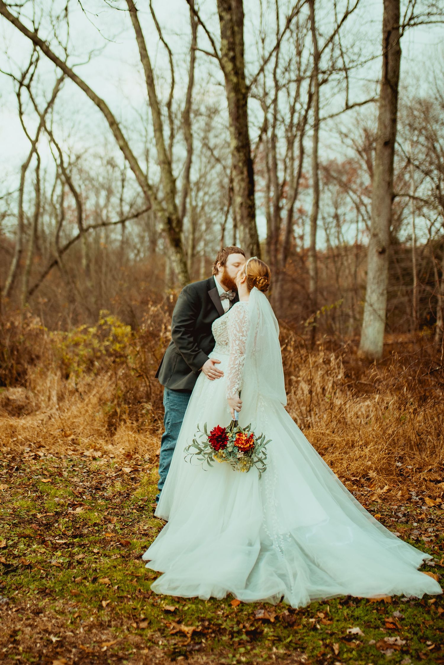 A romantic wedding couple embraces in a rustic autumn forest setting with a flowing white dress and red bouquet.