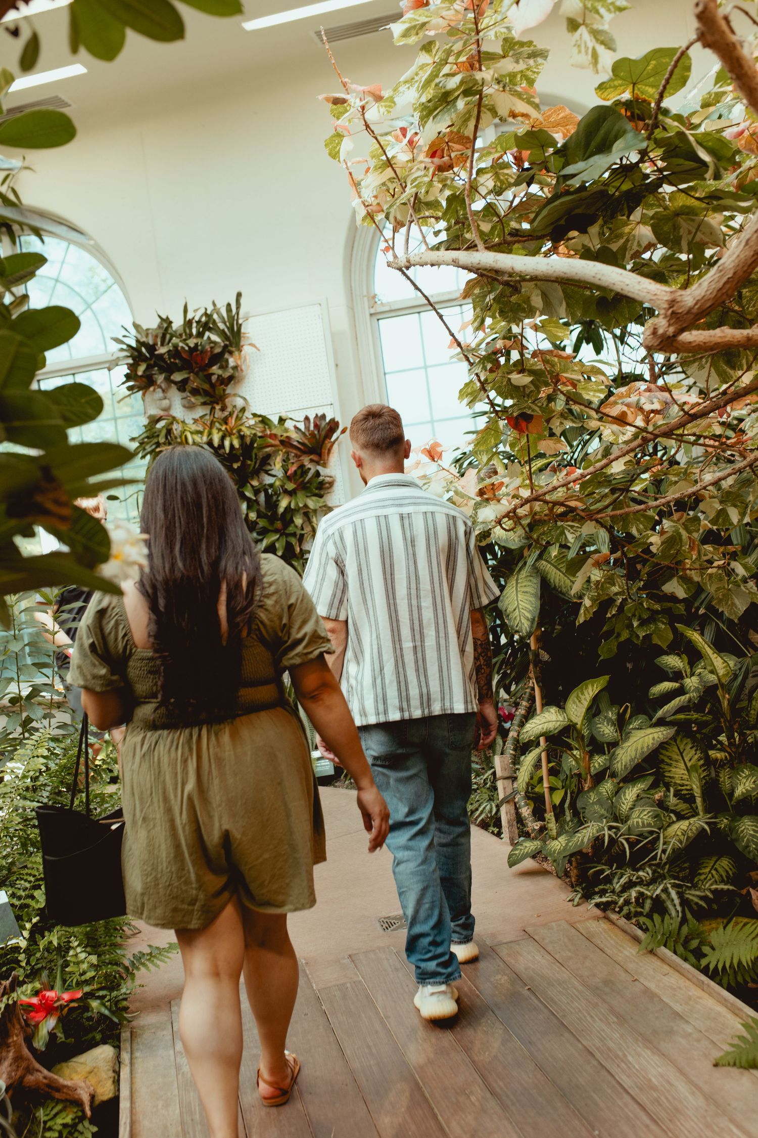 Couple walks through lush botanical garden greenhouse surrounded by tropical plants on wooden path.