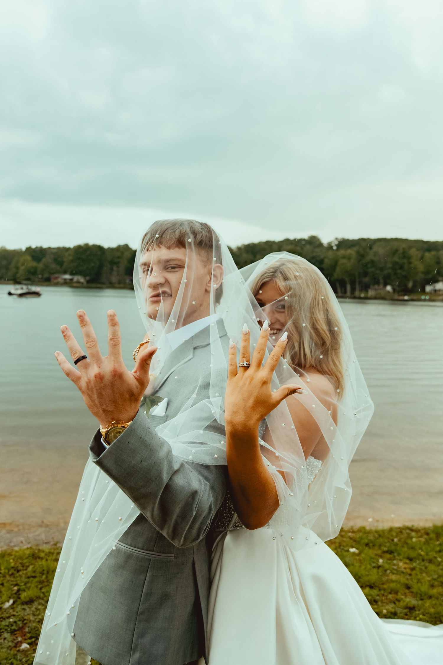 Bride and groom showing wedding rings while sharing veil at lakeside wedding venue.