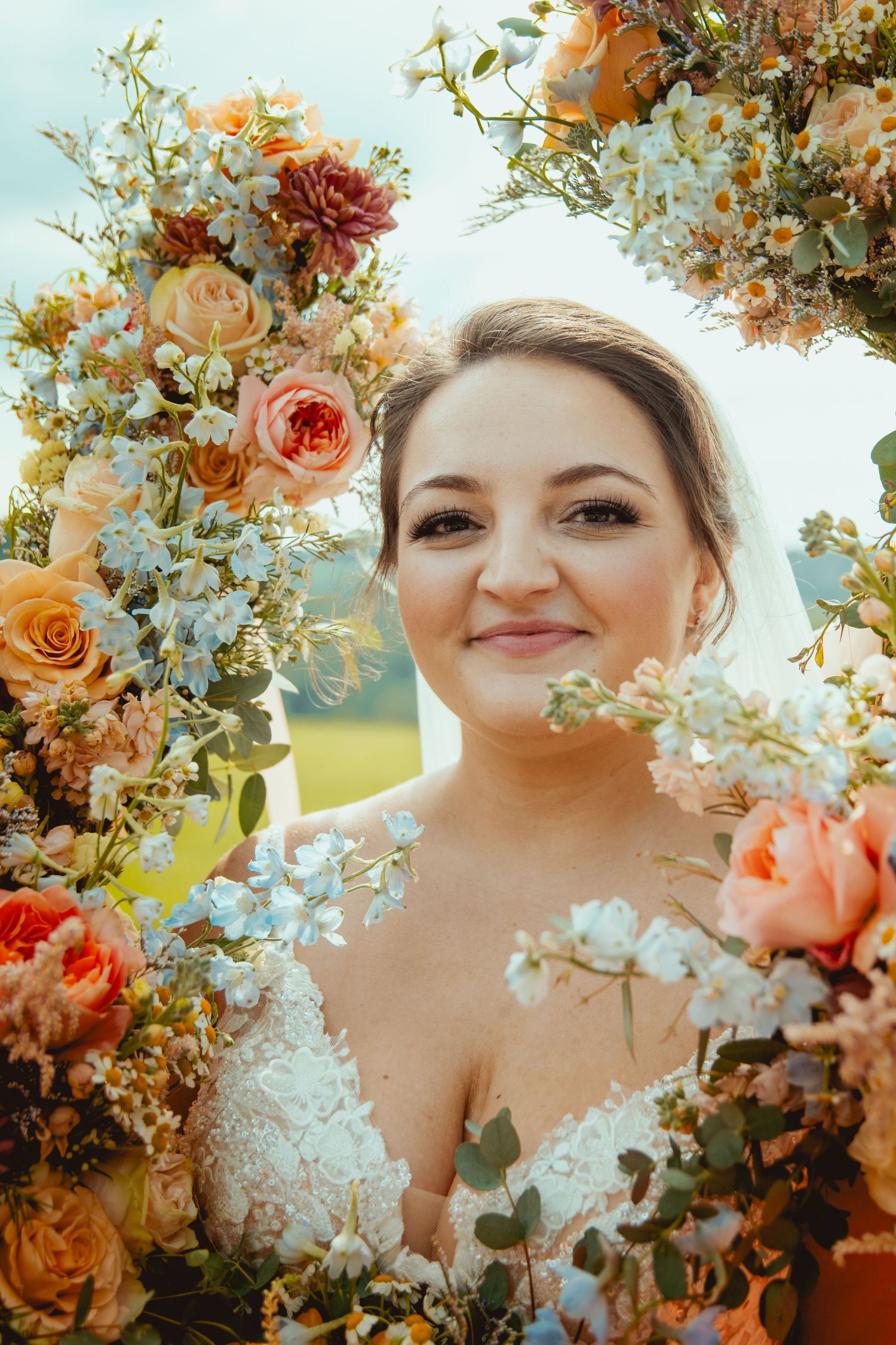 Smiling bride surrounded by lush orange, peach, and blue floral arrangements during an outdoor wedding ceremony.