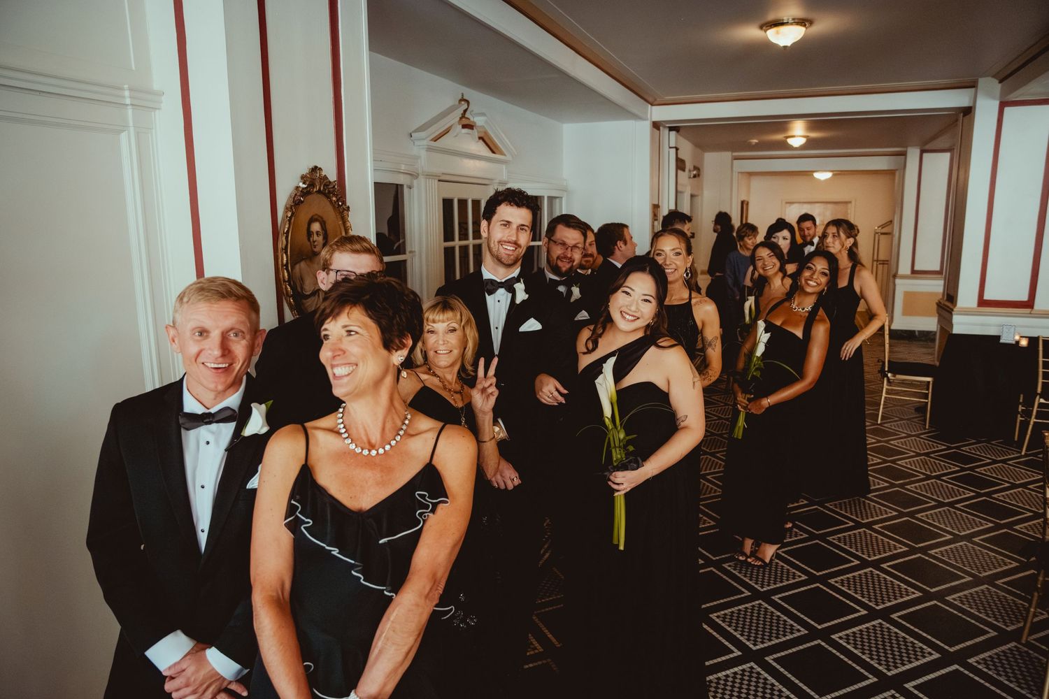 Wedding guests in formal attire gather in an elegant ballroom with patterned carpet and chandeliers.