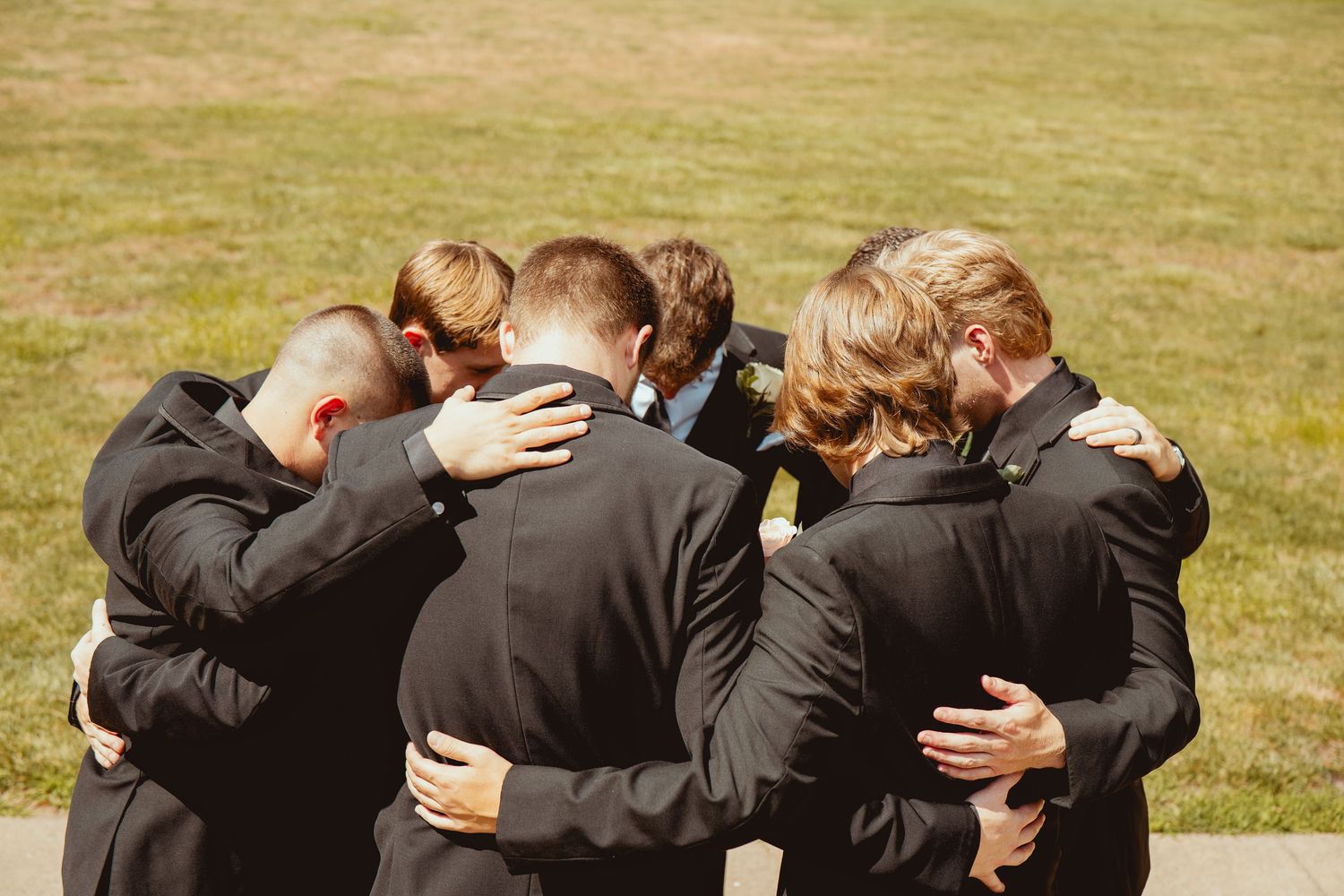 Wedding party members stand close together in a line on grassy lawn during outdoor photo session.