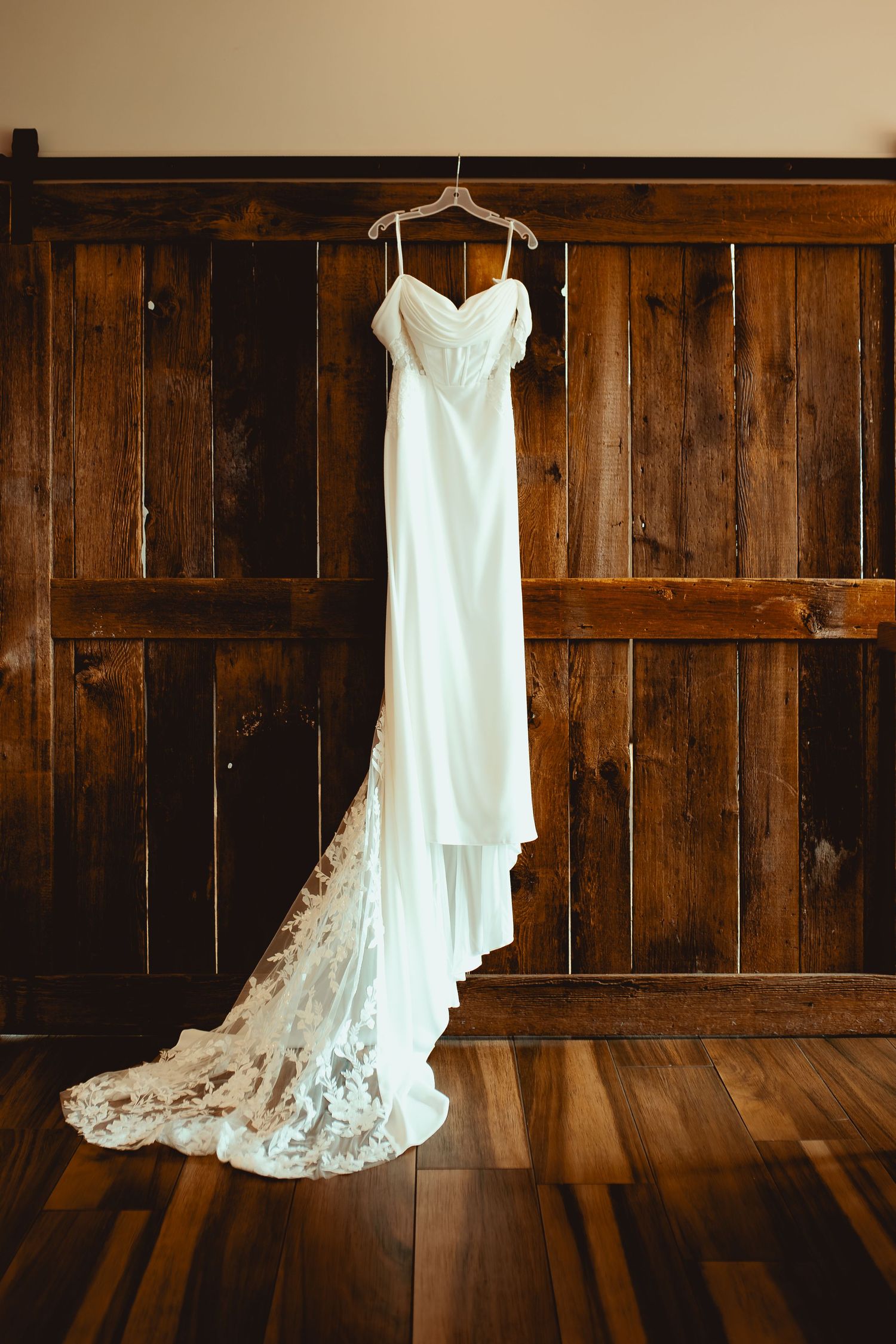 Elegant white wedding dress with lace train hanging on wooden wall, showcasing sweetheart neckline and spaghetti straps.