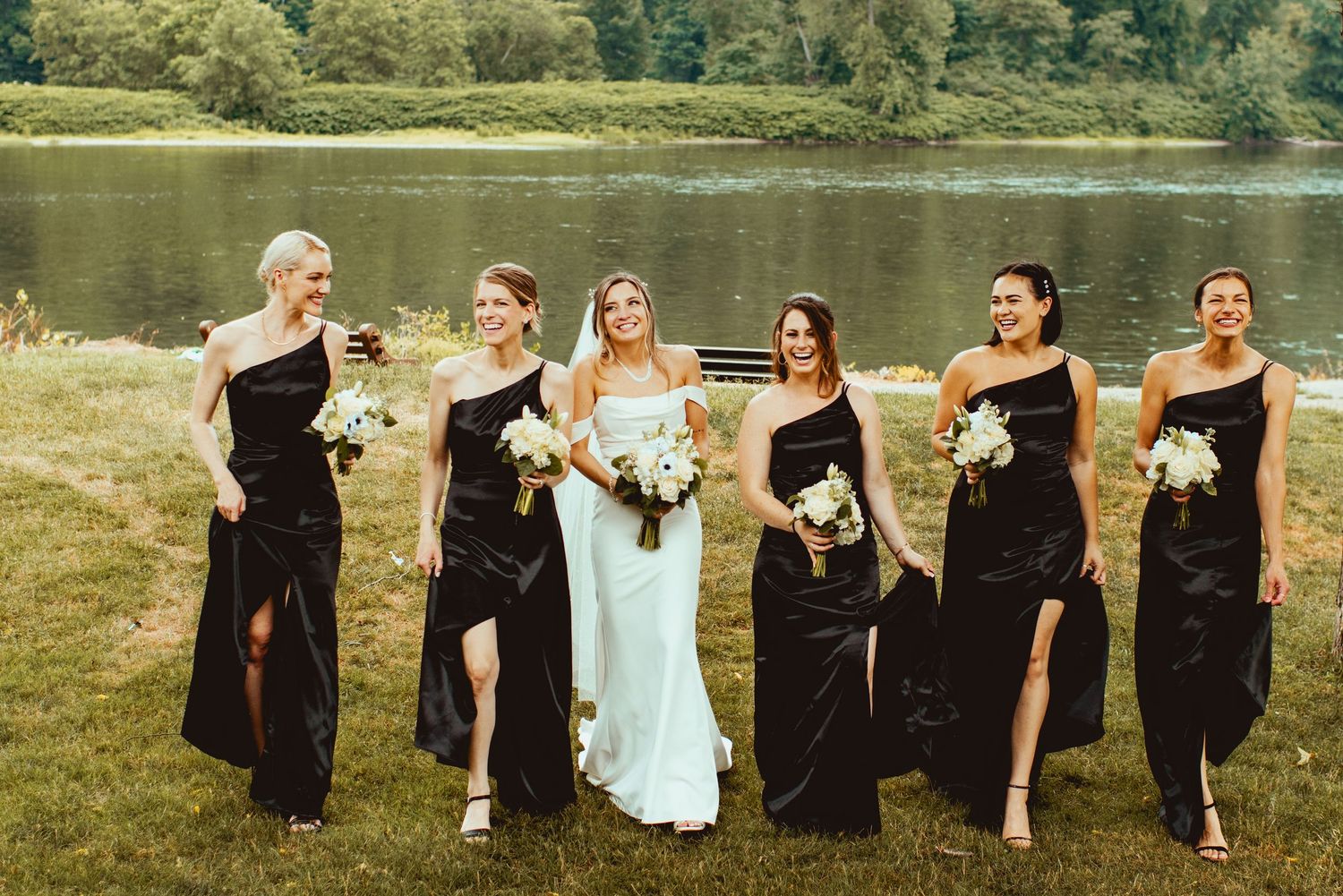 Wedding party posing by a lake with bridesmaids in black dresses and scenic natural backdrop.