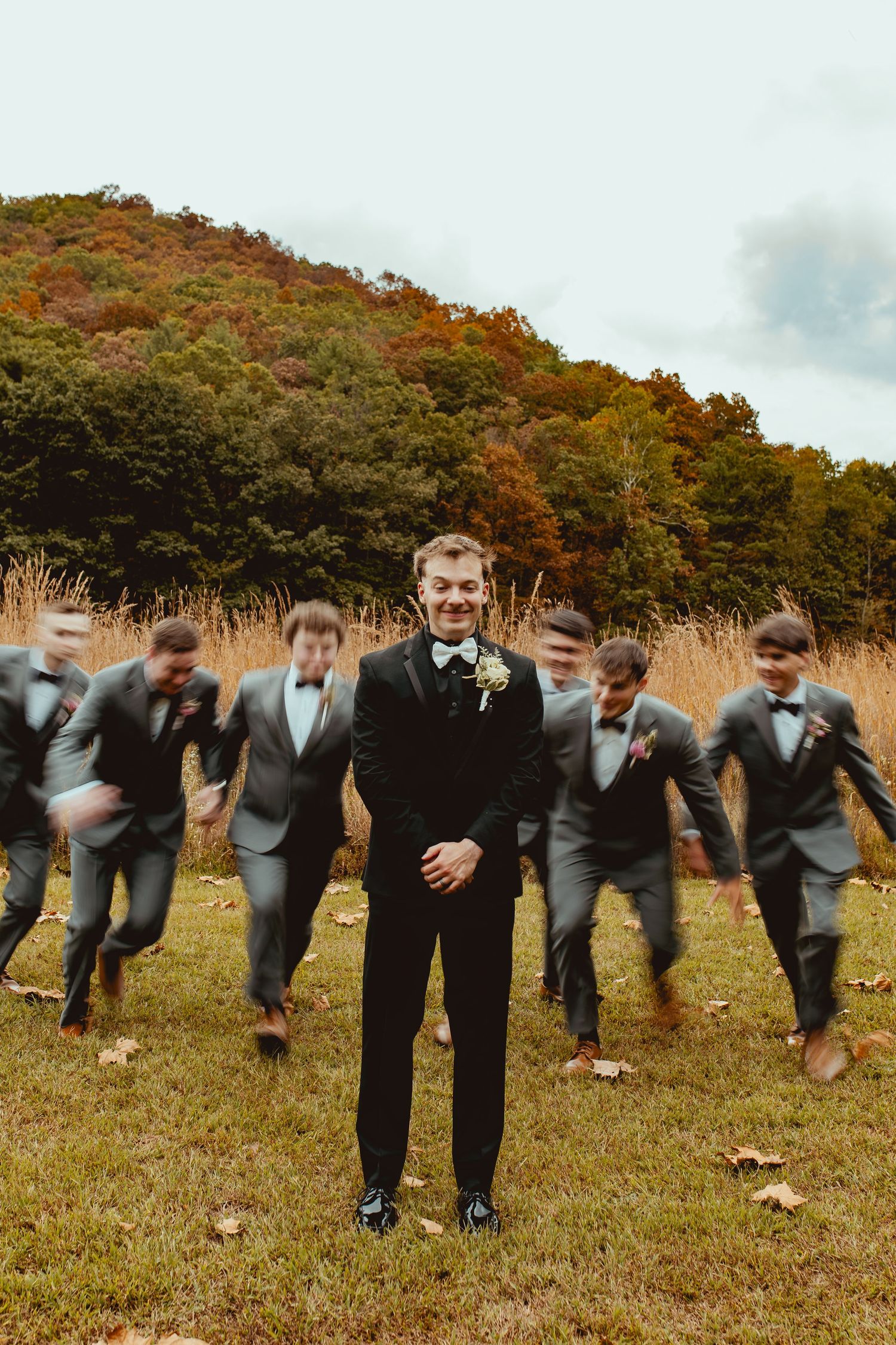 Groomsmen in gray suits perform a synchronized falling domino effect pose on grass with autumn trees in background.