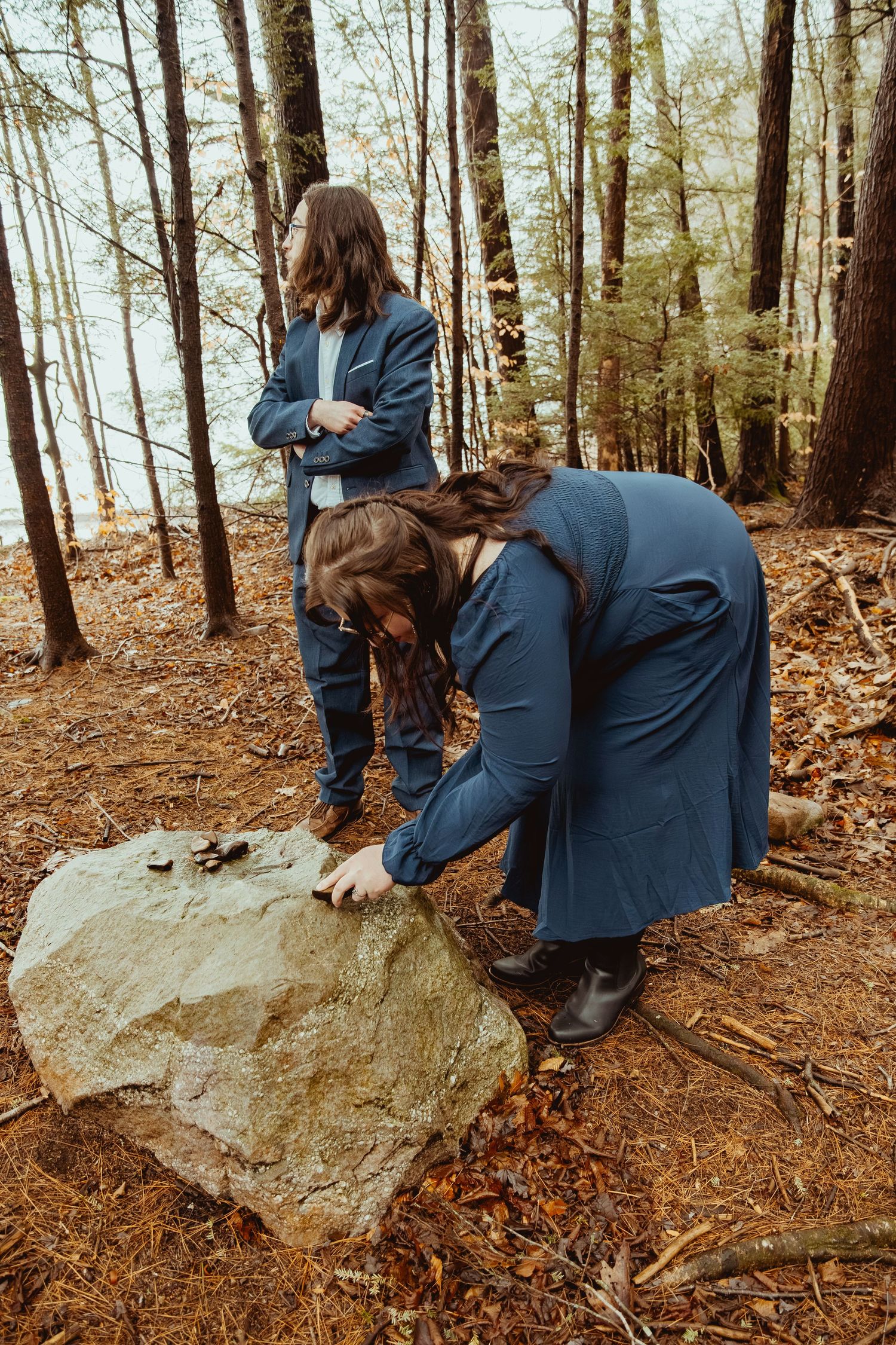 Two people in blue jackets explore and climb on large rocks in a wooded forest setting.