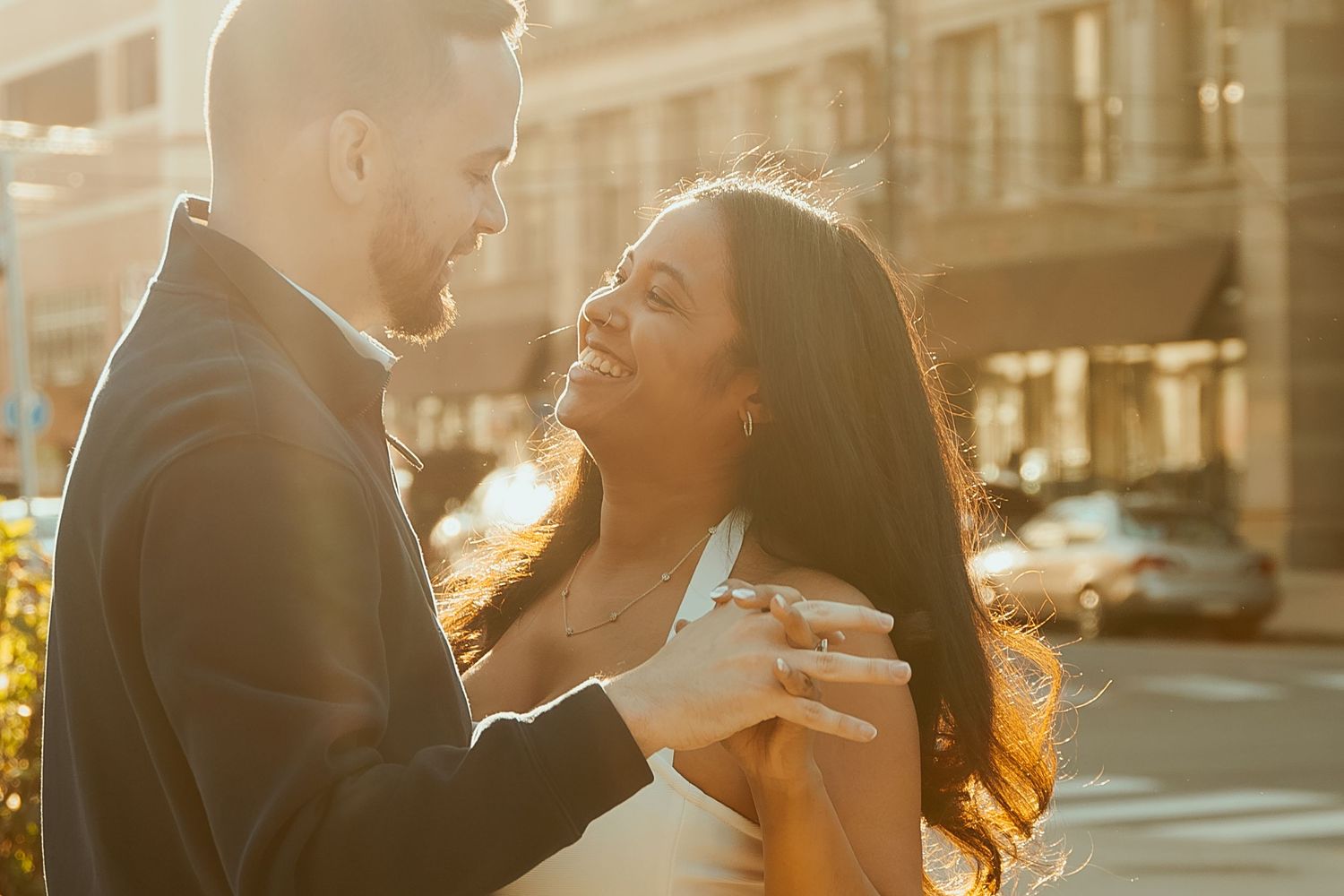 Couple laughing together outdoors at sunset with warm golden light and city buildings in background.