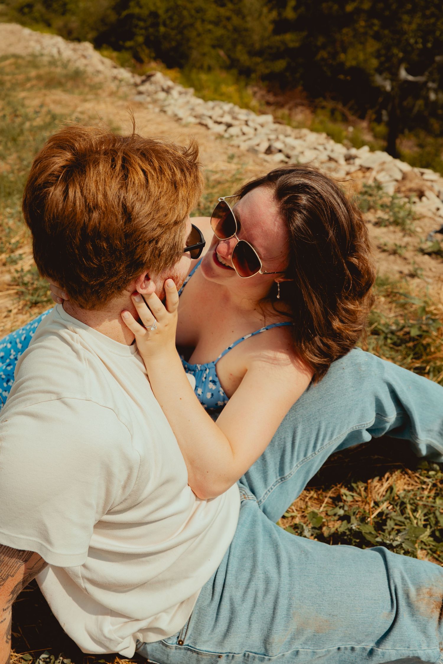 A romantic couple in sunglasses share an intimate moment while sitting on a grassy hillside on a sunny day.