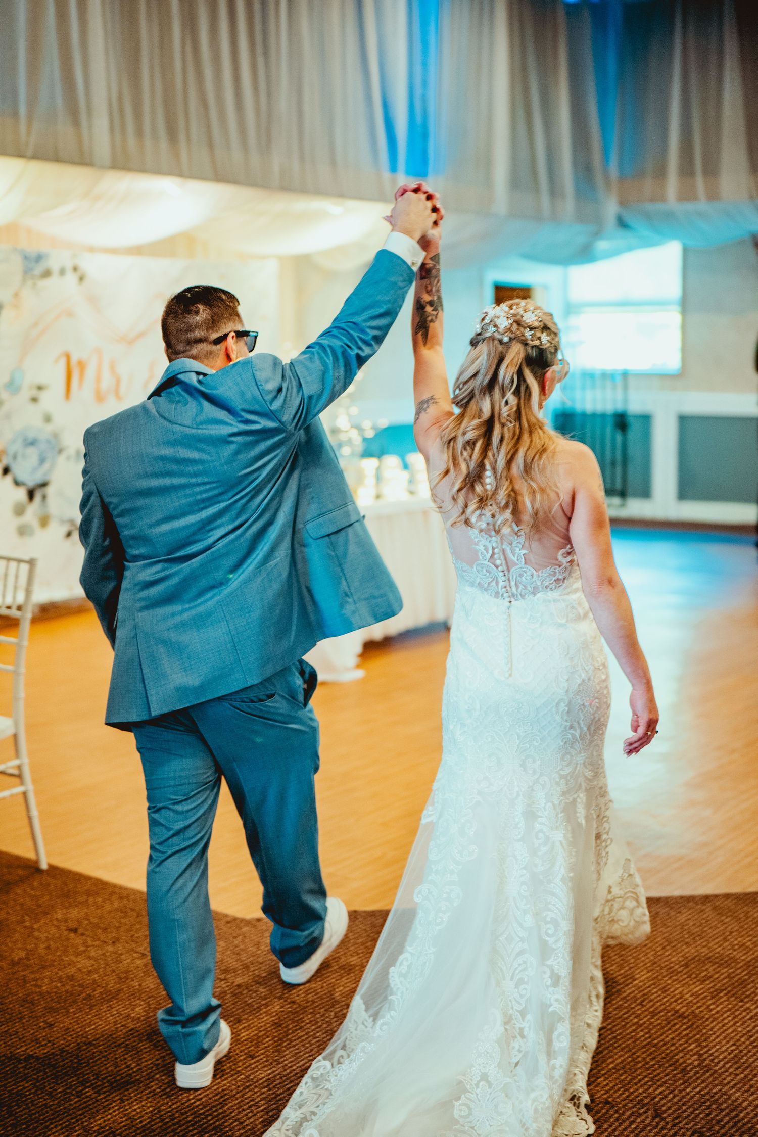 Couple dancing on wooden floor, bride in lace gown and groom in blue suit.