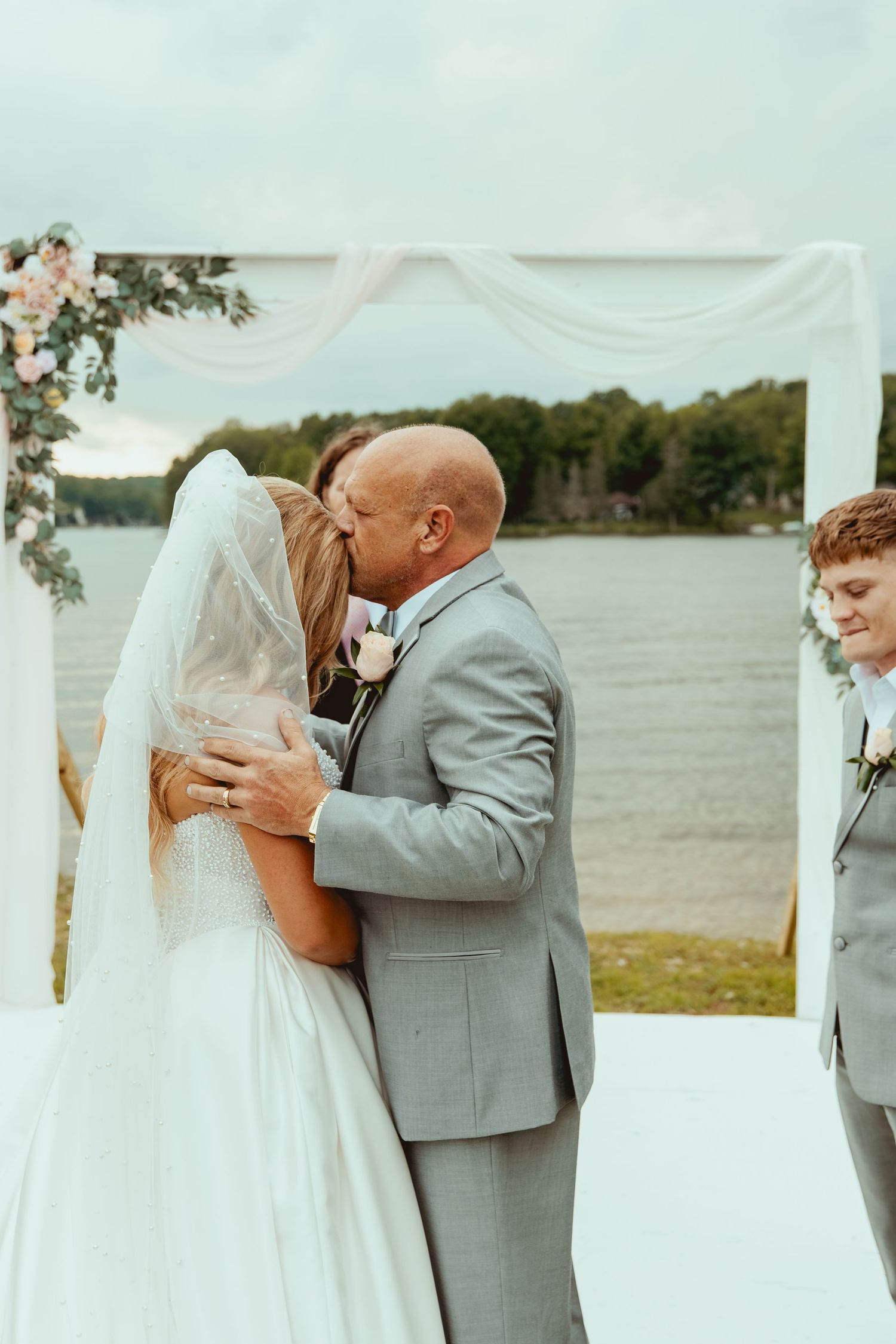 Bride and groom share first kiss at outdoor lakeside wedding ceremony under floral arch.