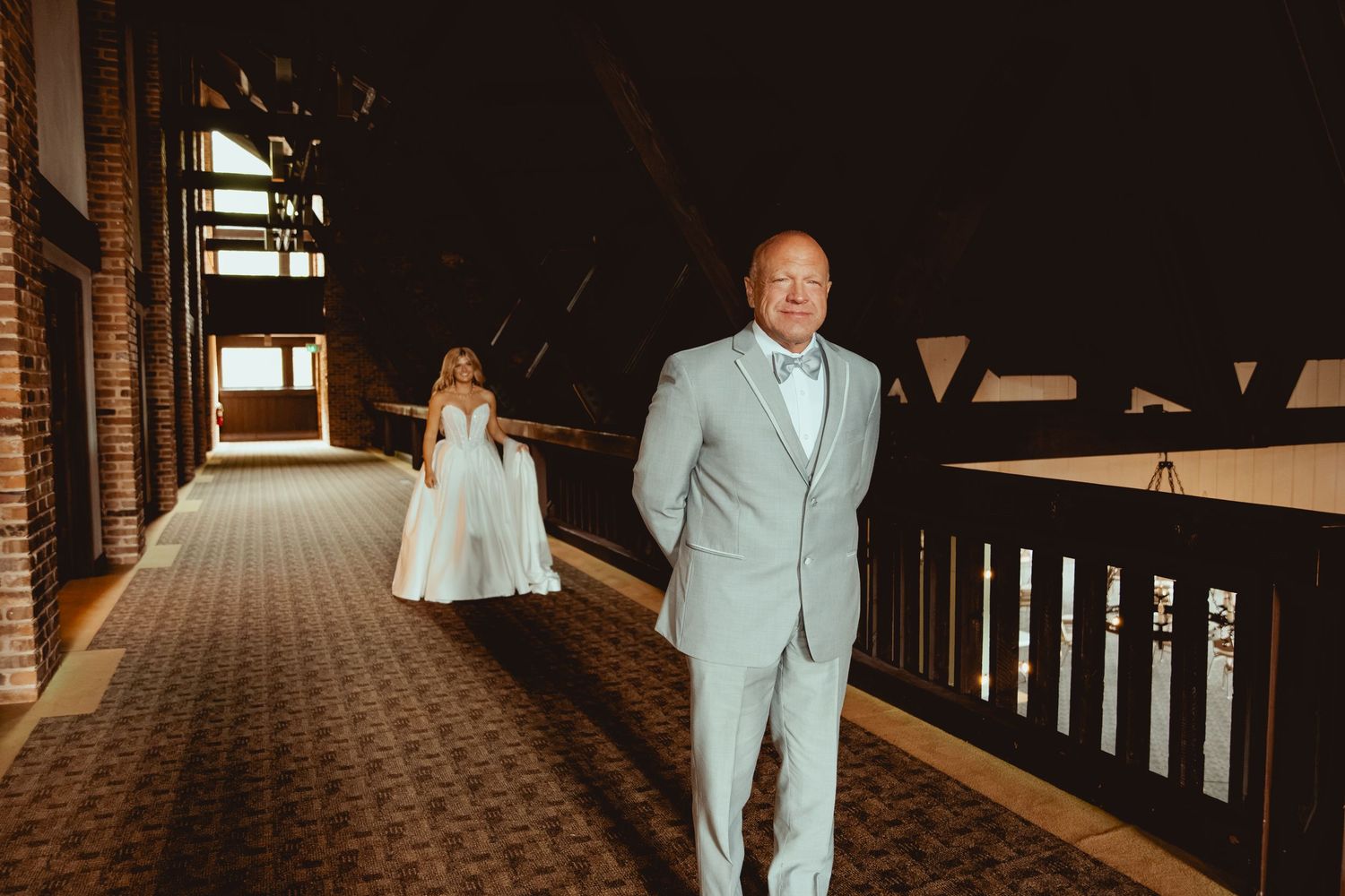 Groom in light gray suit walks ahead of bride in white wedding gown in a warmly lit indoor corridor.
