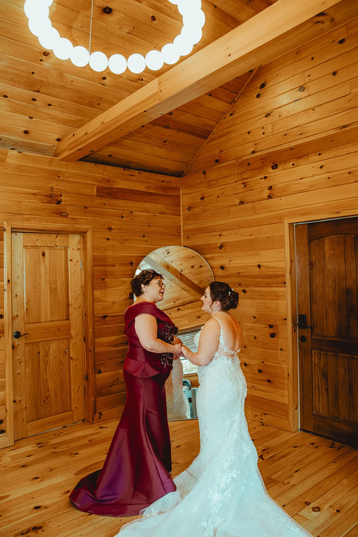 Bride in lace gown getting ready in rustic wood cabin with woman in burgundy dress helping her.