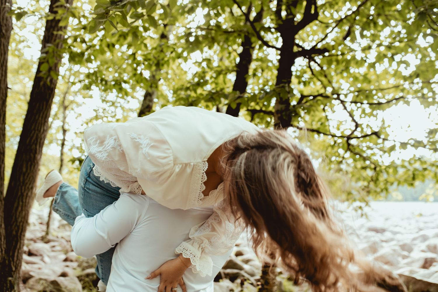 A bride in a white dress being lifted by her partner among lush green trees in an outdoor woodland setting.