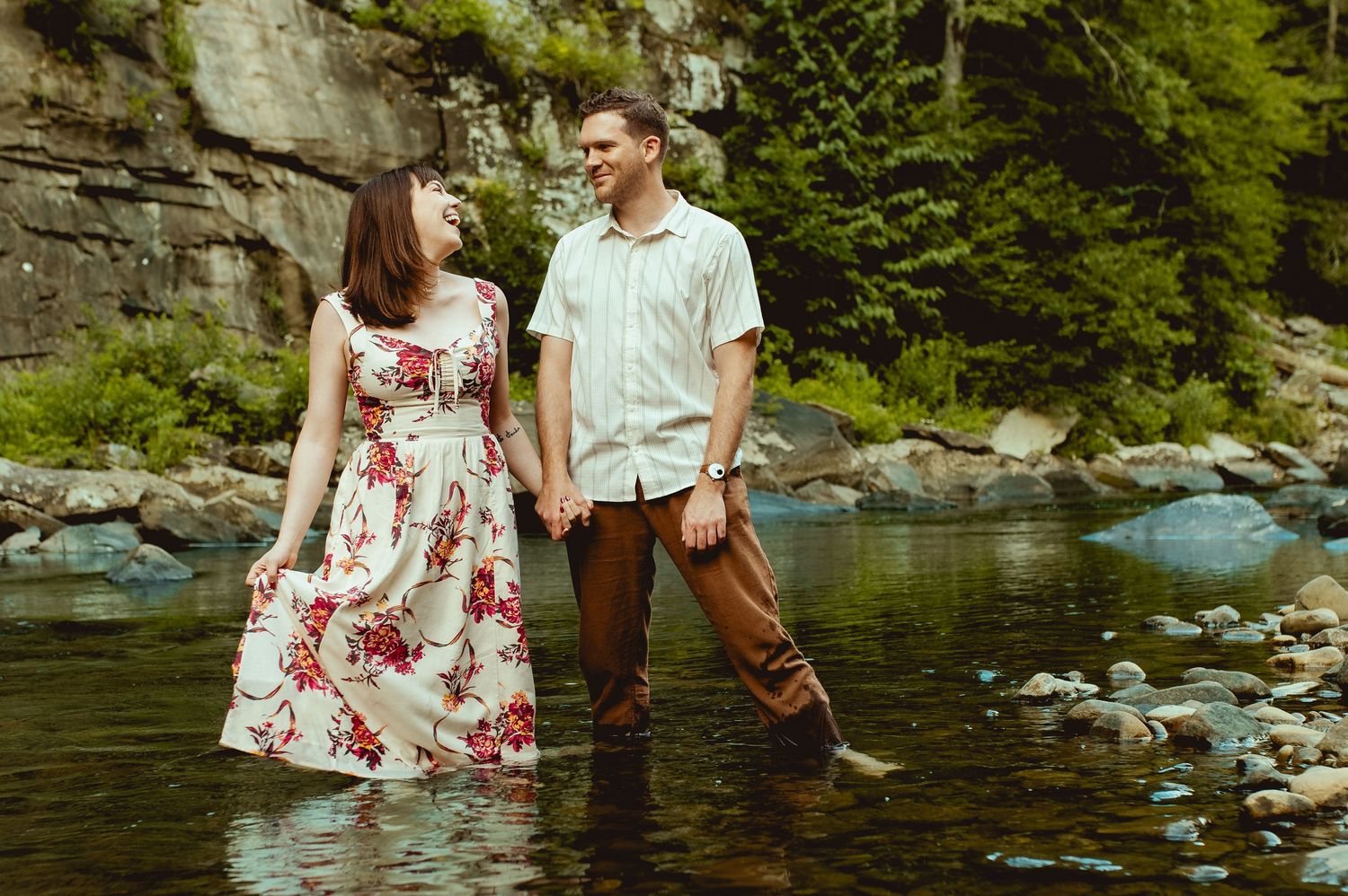 Couple holding hands standing in a rocky river, woman in floral dress, man in casual shirt and brown pants, lush greenery.
