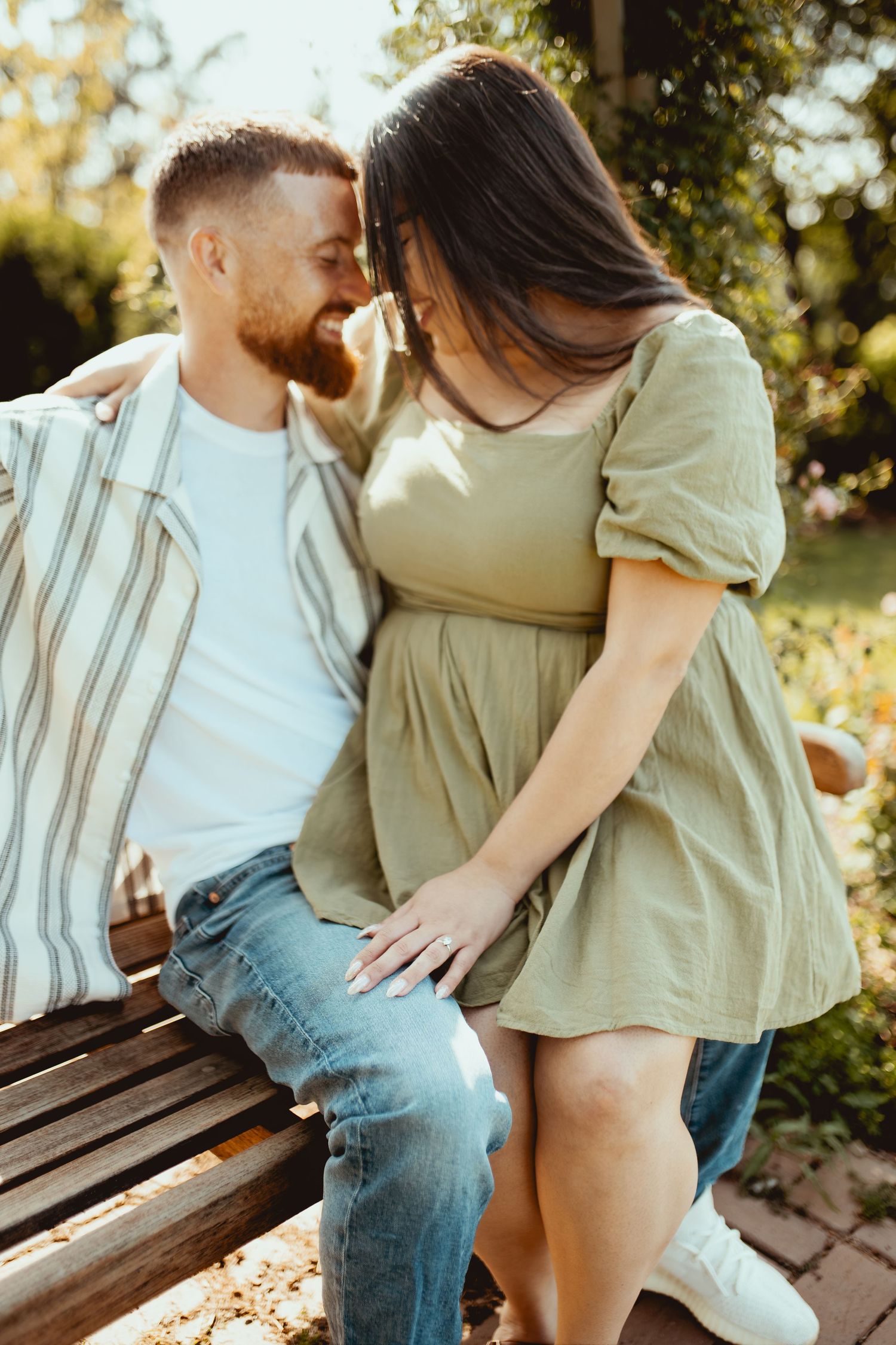 A couple sharing a romantic moment on a park bench, with the woman in a sage green dress sitting on the man's lap.