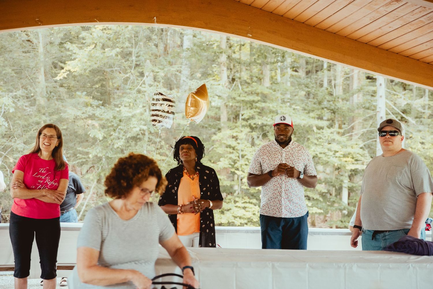 A group gathers under a wooden shelter in a forest setting during a casual outdoor event.