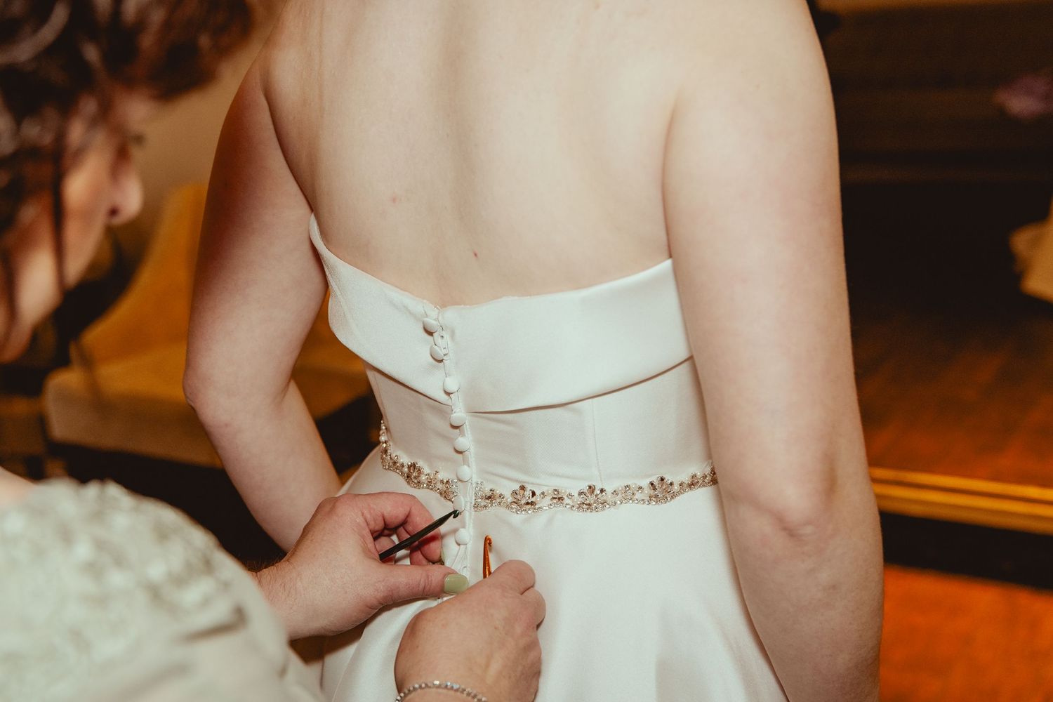 Bride in white strapless wedding gown with pearl buttons and embellished belt being fastened by bridesmaid.