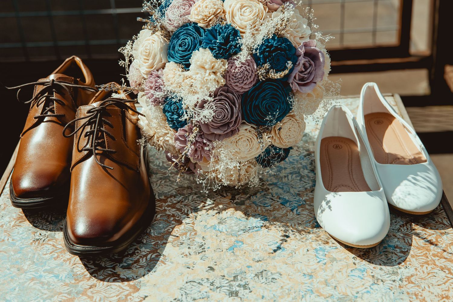 Brown leather dress shoes and white bridal flats beside a teal and pink flower bouquet on a marble surface.