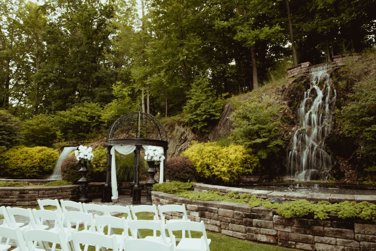 Outdoor wedding ceremony setup with white chairs, floral arch, and scenic waterfall surrounded by lush greenery.