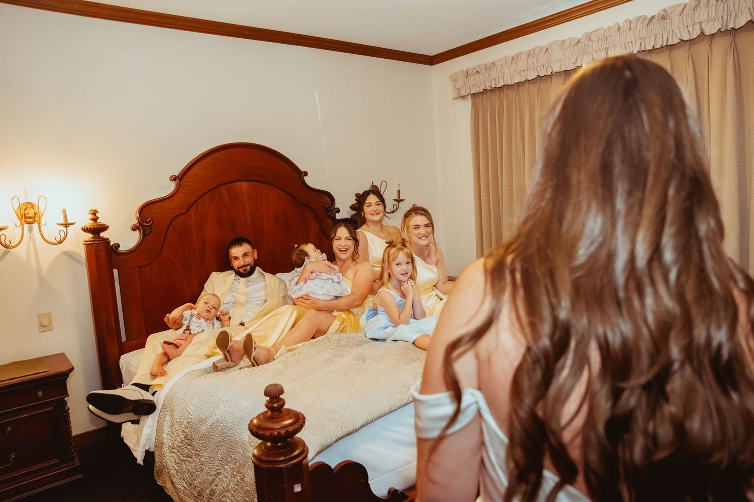 Family gathering on a bed as a bride in pink robe reveals her wedding dress during bridal prep.