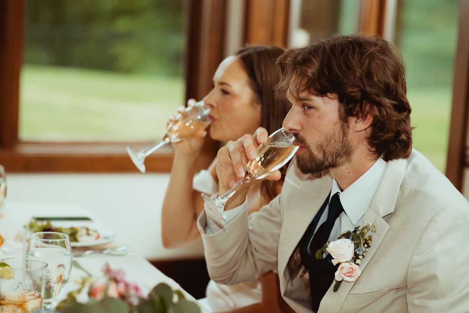 Bride and groom in beige suit sip champagne together at wedding reception dinner table.