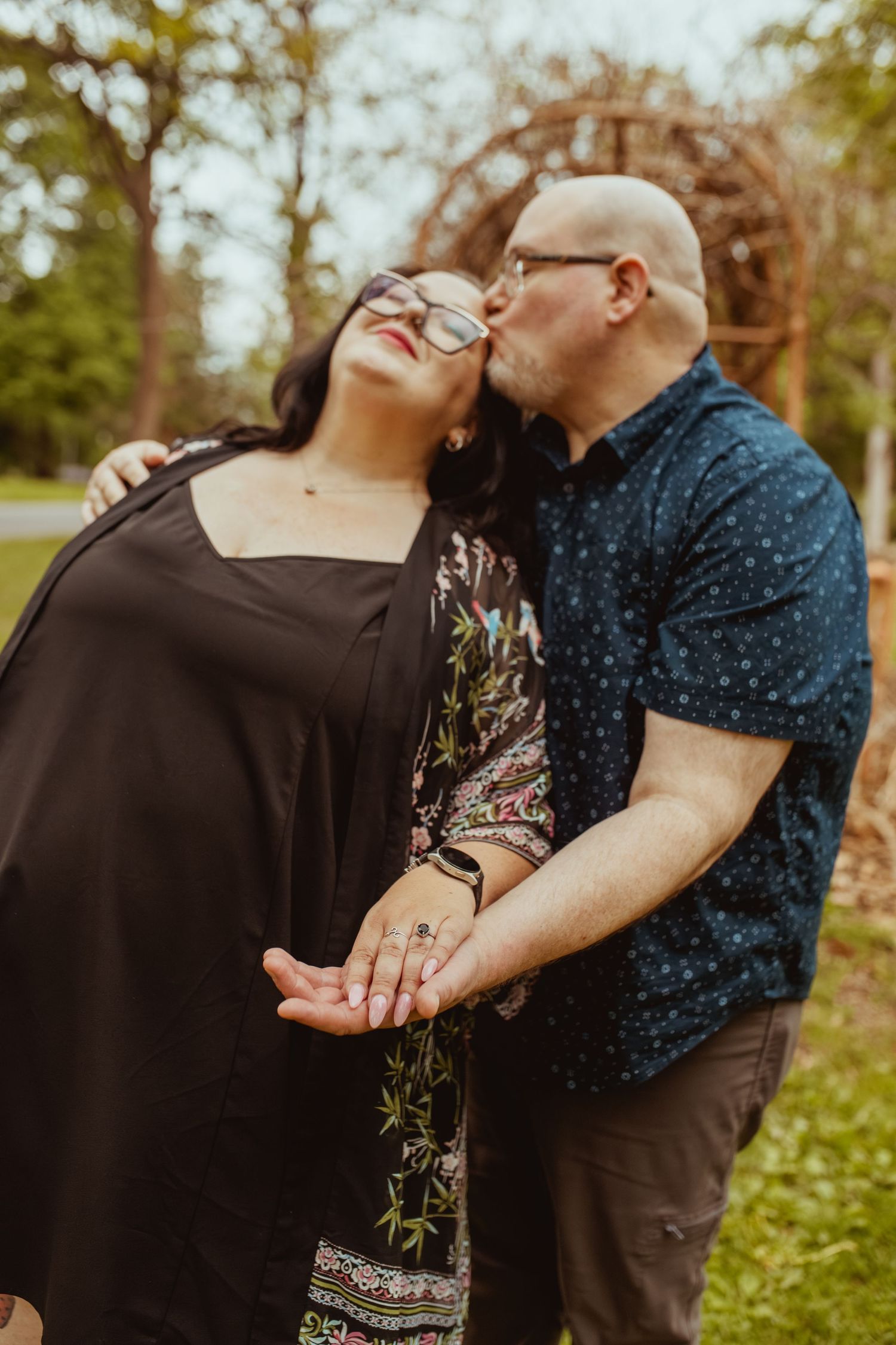 Bald man kissing woman on the cheek as she leans back with eyes closed outdoors.